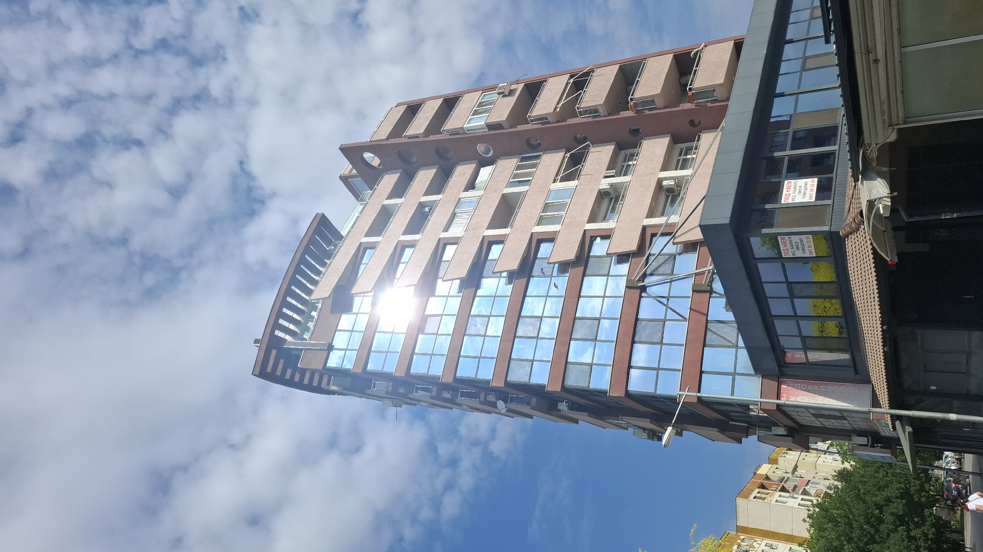 This image captures a tall, modern building with a distinctive curved roofline, situated in the city of Plovdiv, Bulgaria. The building features a facade predominantly composed of reflective blue-tinted windows, interspersed with sections of pale pink and reddish-brown cladding. Numerous balconies with light-colored railings extend from the upper floors. On the lower part of the building, there are shops with large glass windows, one of which displays signage in Bulgarian that reads "ПОД НАЕМ" (FOR RENT) along with phone numbers. A street light and a lamppost are visible near the building's entrance, and trees and other urban structures, including apartment buildings, are present in the background. The sky is partly cloudy with bright sunlight reflecting off the building's windows. A single person is partially visible in the lower-left corner of the image, walking away from the camera.