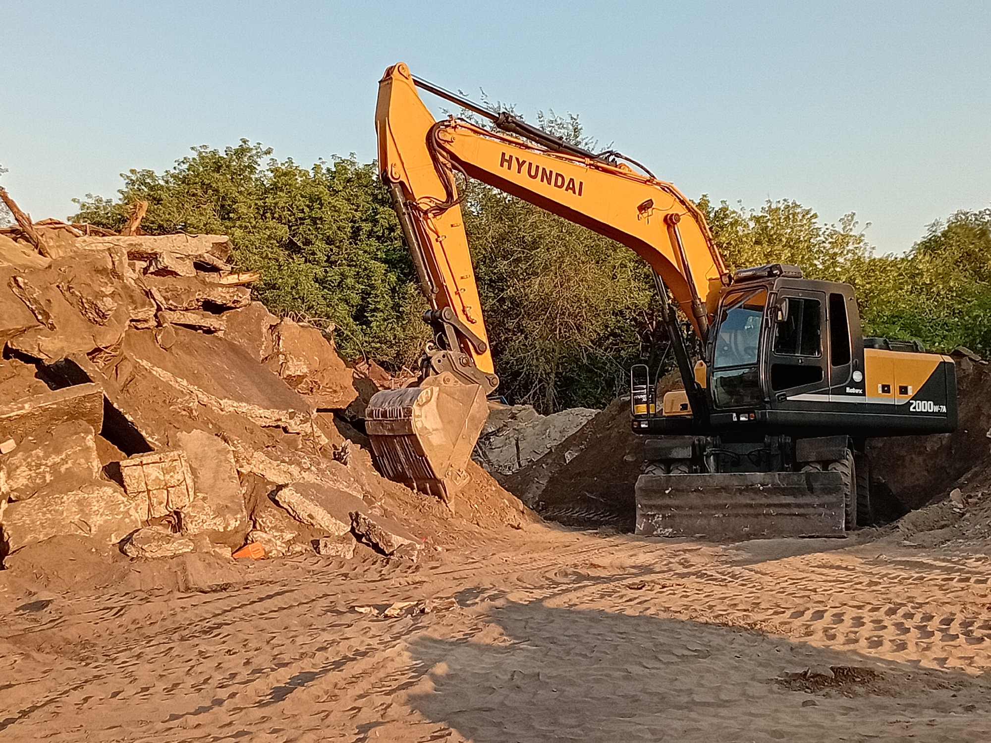 An excavator with "HYUNDAI" visible on its boom is positioned in a construction or demolition site. The excavator is yellow and black and has the model number "2000W-7A" on its side. In the foreground, there is a pile of large concrete debris, and the ground is covered in sand with tire tracks. Behind the excavator and debris, there is a line of green trees. The sky is clear and light blue, suggesting it is daytime. The scene appears to be an outdoor area, possibly on the outskirts of a city.