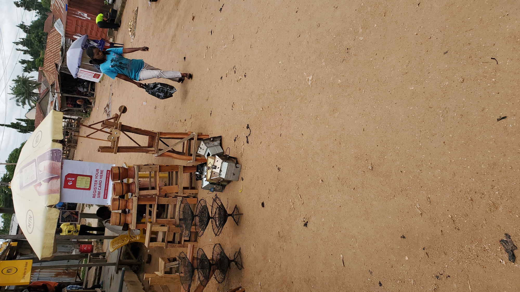 A street market scene in Jalingo, Nigeria, features a woman in a blue t-shirt and white pants walking with a black plastic bag. She is the primary human subject in the foreground. In the background, a market stall displays wooden bowls on a wooden stand, with a large umbrella overhead. A sign on the stall reads "REGISTER YOUR SIM CARD HERE". To the right of the stall are stacked metal components, possibly electronic waste or parts. Further in the background, other market stalls with corrugated iron roofs are visible, and several individuals are present, including a person in green in the far background. The ground is sandy and appears to be a dirt road. The overall atmosphere suggests a bustling but somewhat unorganized marketplace.