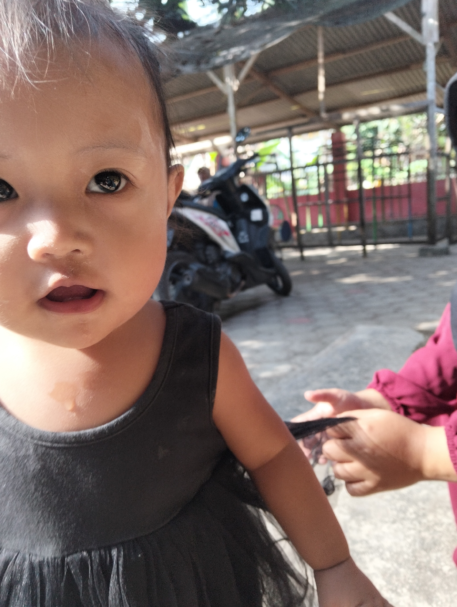 A close-up shot focuses on the face of a young child. The child has dark eyes and an open mouth. The child is wearing a dark grey dress. To the right of the child, another person’s hands are visible, holding a dark, wispy material. In the blurred background, a motorcycle is parked under a covered outdoor structure with a tin roof, supported by wooden beams. Beyond the structure, a red fence and green foliage are visible. The ground appears to be paved with concrete or stone. The overall scene suggests an outdoor, possibly communal or public, area in a village setting. The image is taken outdoors in what appears to be a village in Desa Wringin Putih, Indonesia.