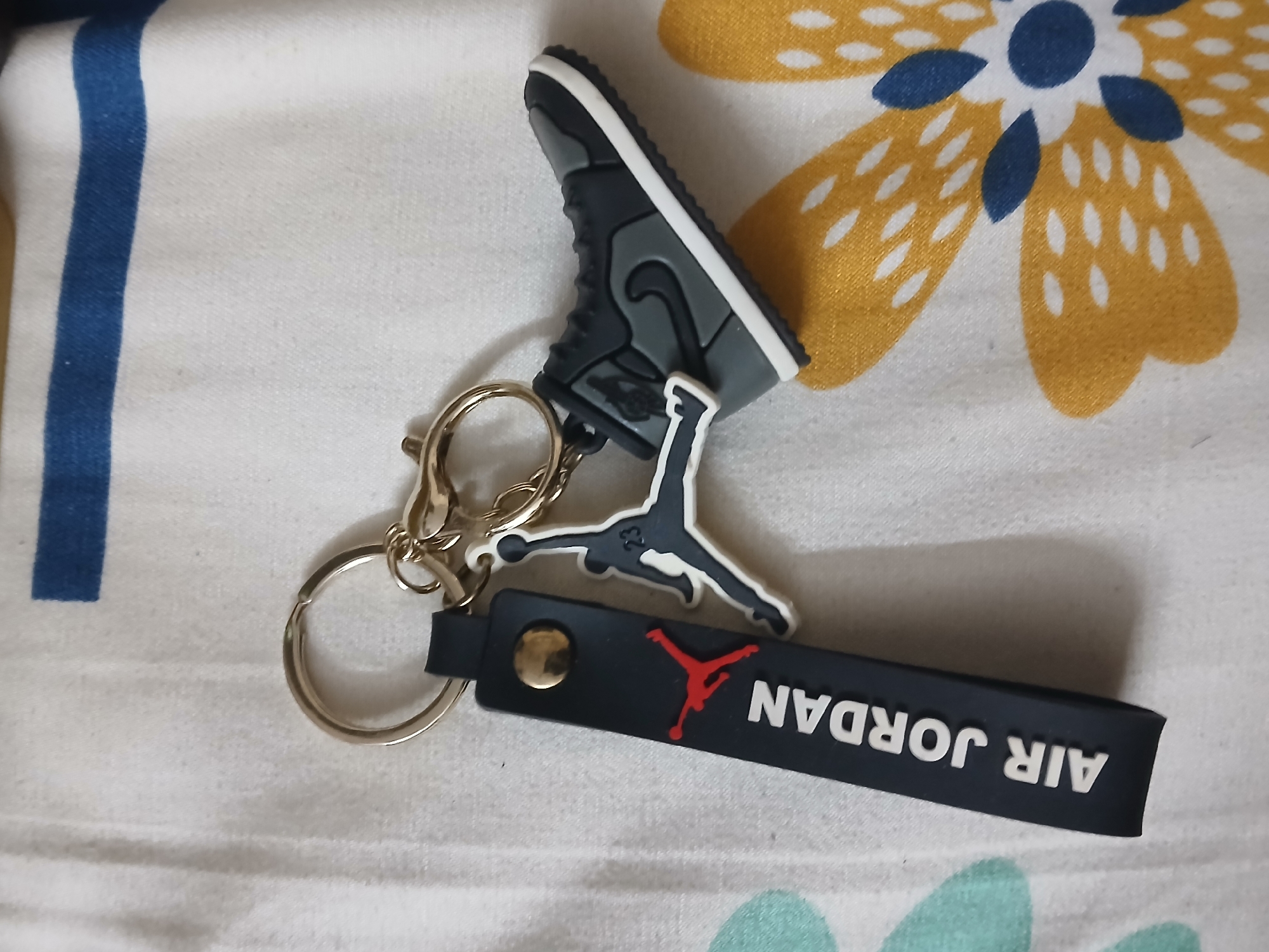 A close-up shot presents an Air Jordan keychain on a patterned fabric surface. The keychain features a miniature sneaker, the iconic Jumpman logo, and a black rectangular tag labeled "AIR JORDAN." The background showcases a flower pattern in yellow, white, and blue. The fabric seems to be part of bed linen. Given the prompt, this image could be taken anywhere in Bengaluru, India.