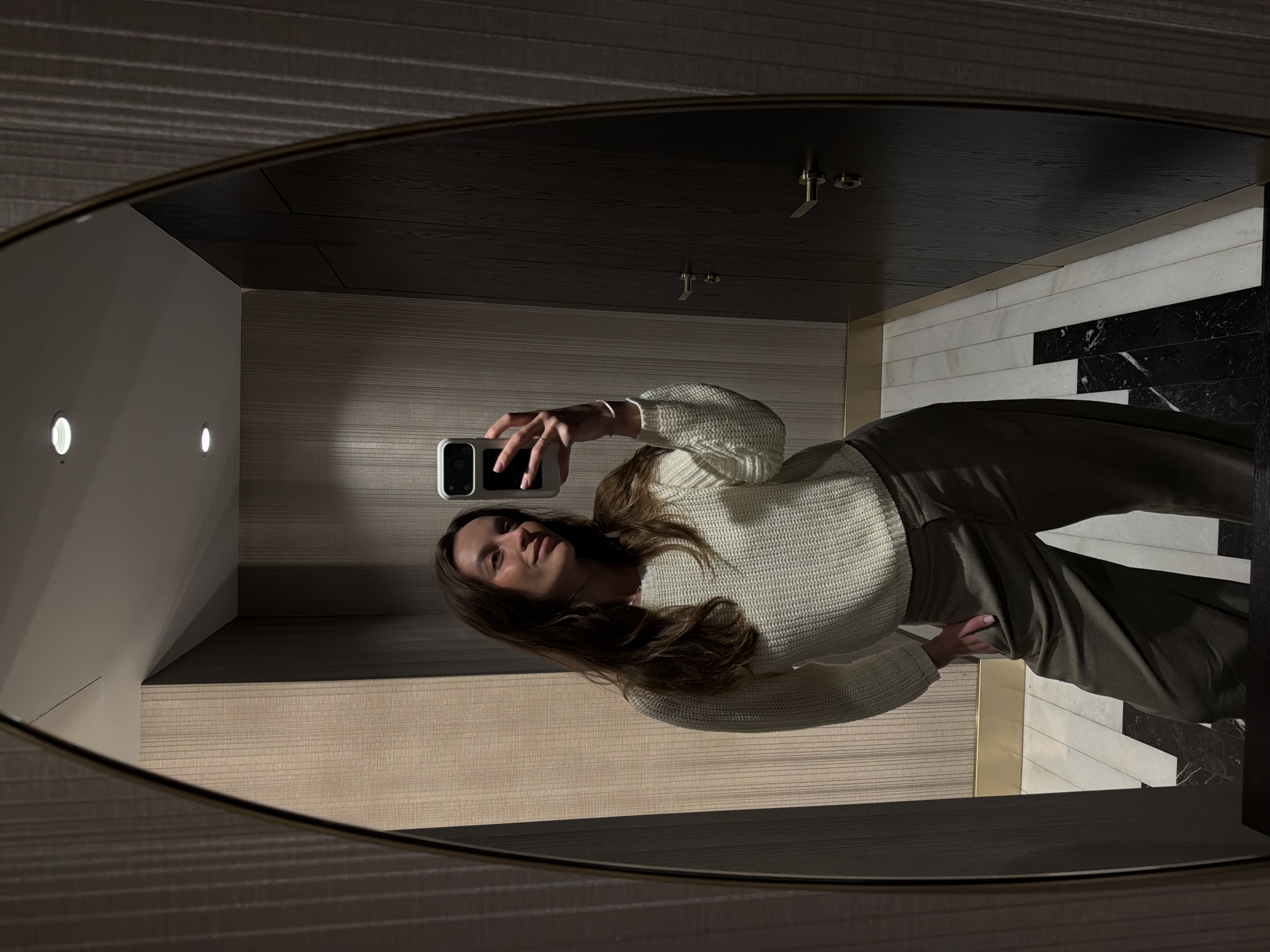 A woman is taking a selfie in a mirror in an interior space. The space features a light-colored paneled wall with two embedded lights, dark-stained wooden paneling on the ceiling, and a black-and-white marble-patterned wall. The woman is wearing a white sweater and olive-green pants. The location is likely a bathroom or dressing room, but the city of Budva, Montenegro, cannot be determined from the image.