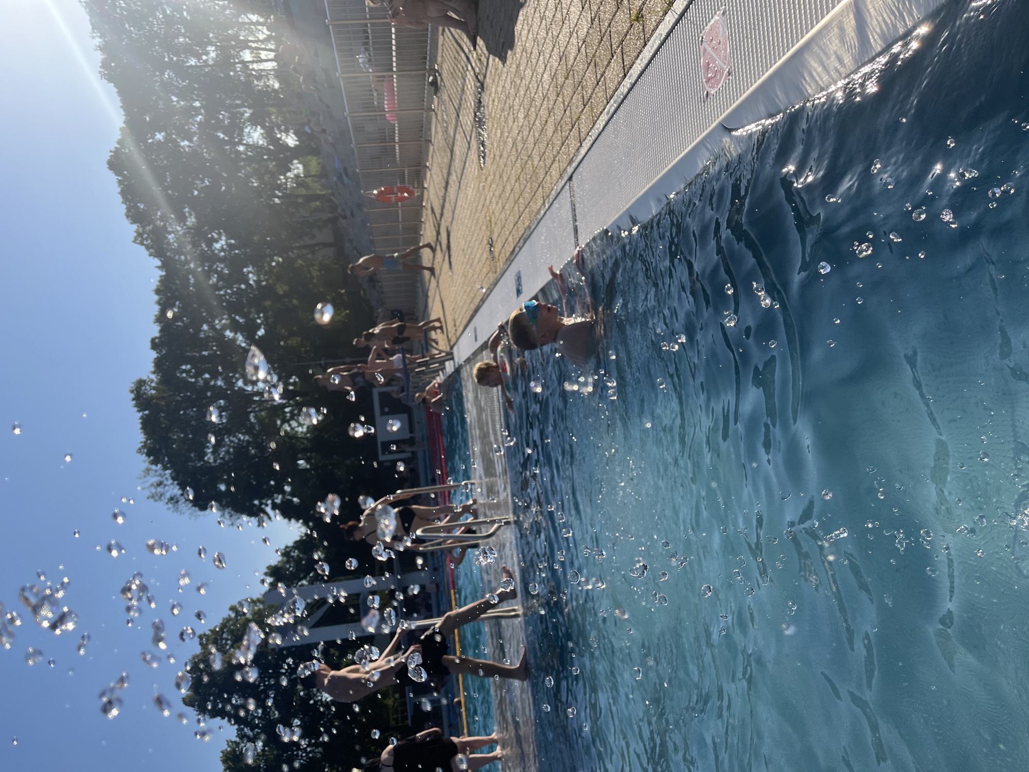 This media file depicts a group of people, primarily children, at an outdoor swimming pool on a sunny day. The image is captured from a low angle, looking up at the sky and across the pool. Many of the individuals are in various stages of jumping or preparing to jump into the water from a raised platform or diving board, indicated by their body positions and the spray of water in the foreground.
The scene is set at a pool with a tiled edge and a metal railing, suggesting a public or recreational facility. Trees and a clear blue sky are visible in the background, reinforcing the outdoor setting and warm weather. The sunlight is bright, creating glares on the water's surface and casting long shadows. The overall mood seems to be one of fun and excitement, with the splashing water adding to the dynamic nature of the activity.
Notable details include the presence of goggles on at least one individual and a life preserver visible on the side of the pool. There are also several water droplets captured in mid-air, adding a sense of motion and energy to the image. The time of day appears to be daytime, likely late morning or afternoon, due to the bright sunlight and the angle of the shadows. No discernible text is visible in the image.