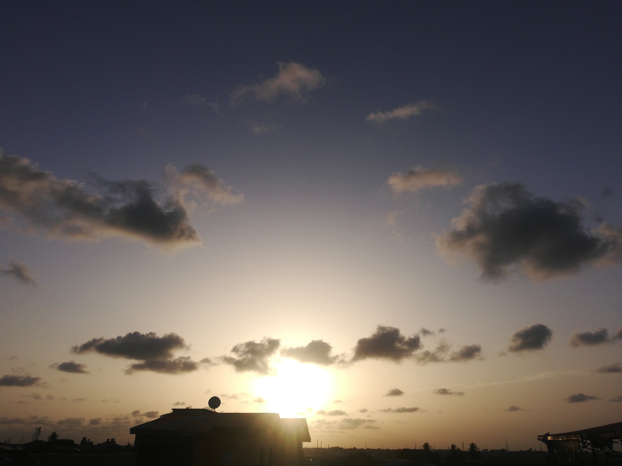 The image depicts a low-angle view of a sunset in Lagos, Nigeria. The bright sun is positioned near the horizon, partially obscured by clouds. A dark silhouette of a building with a satellite dish on its roof is prominent in the foreground. The sky transitions from a lighter hue near the horizon to a darker blue-purple shade in the upper portion of the image. Scattered clouds are visible throughout the sky. Other buildings and trees are visible in the distance along the horizon line.