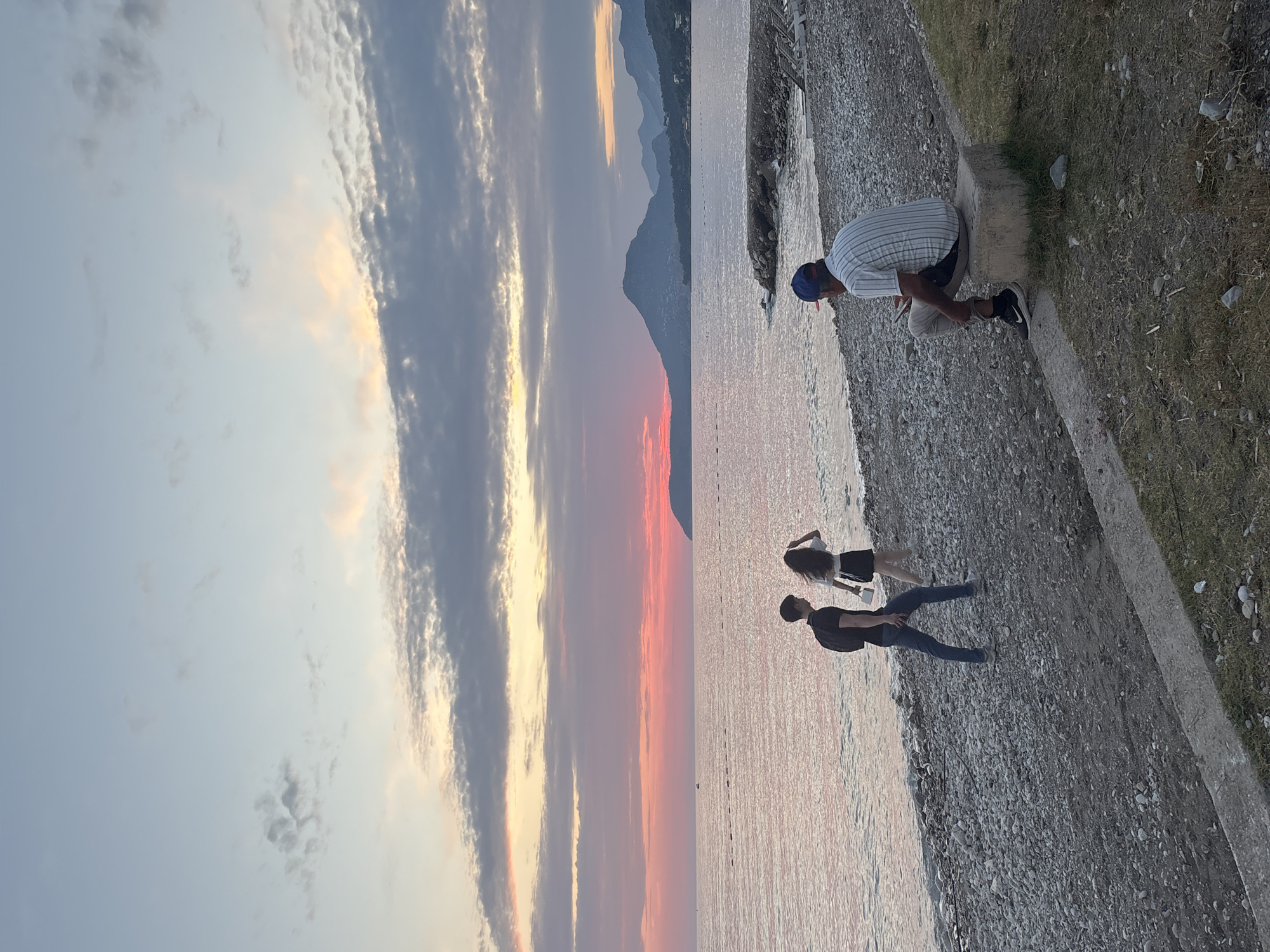 The image depicts a group of people by the sea at dusk. The sky is filled with dramatic clouds in hues of orange, pink, and blue, suggesting a sunset. In the foreground, a man in a striped shirt and blue cap is crouching on a concrete structure near the water's edge, looking down. Further along the shore, two figures, a woman in a white top and black skirt, and a man in a dark shirt and jeans, are standing on a pebble beach. The water is calm and reflects the colors of the sky. In the background, mountains are silhouetted against the twilight. The scene evokes a sense of tranquility and the beauty of nature. There is no visible text in the image. The location context is Bar, Montenegro.