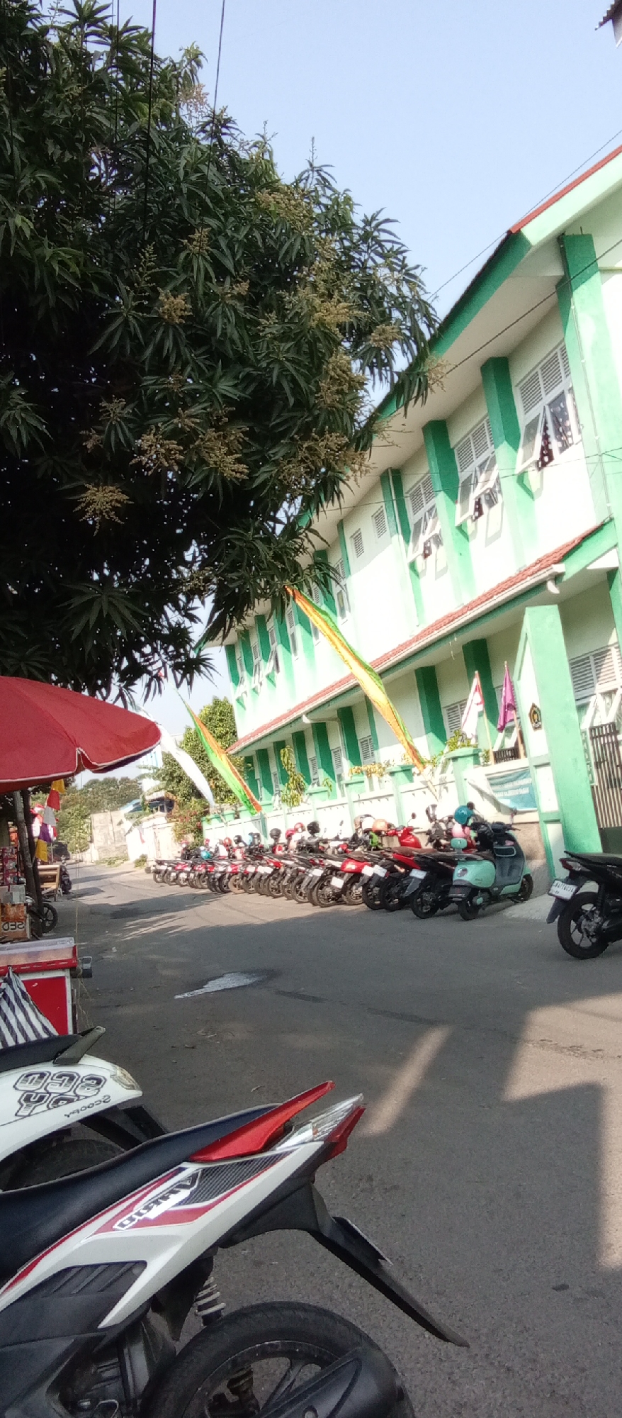In Kediri City, Indonesia, a street scene is depicted with a building and parked motorcycles. A two-story building, painted white with green trim, stands prominently on the right side of the image. The building has numerous windows with white shutters, and some windows are open, revealing glimpses of the interior. Colorful flags are strung across the facade, adding a festive touch. In front of the building, a row of motorcycles is parked along the side of the road, suggesting a public space or a school/office environment. A tree with lush green foliage and small, light-colored blossoms or fruits overhangs the scene from the left, providing shade. In the foreground, the rear section of a white and red motorcycle is visible, with its tire and part of the bodywork in focus. To the left, a red umbrella is set up, possibly for a street vendor, with some goods displayed beneath it. The ground is paved asphalt, and shadows from the tree and the building are cast across it, indicating a sunny day. The sky is clear and blue. The overall impression is one of a typical urban street in Indonesia, bustling with activity and local elements.
