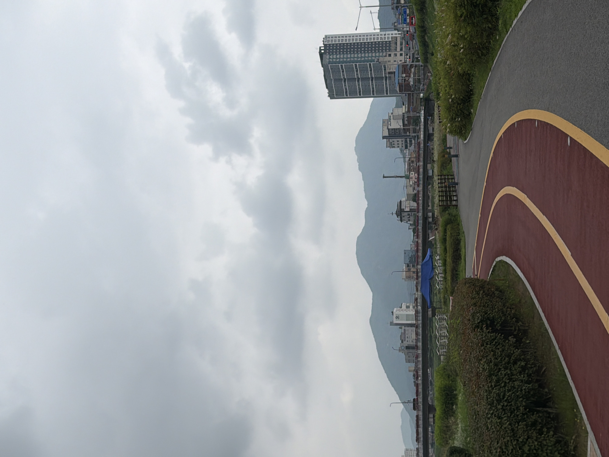 This is a vertical, eye-level, medium shot of a paved park path with red and yellow markings, curving from the bottom left towards the center right of the frame. A dense, green hedge forms the left edge of the path. In the background, a city skyline with various buildings, including a tall, multi-story building on the right, is visible against a backdrop of soft, rolling mountains under a cloudy sky. A bridge spans a waterway in the mid-ground. A blue tarp is visible beneath the bridge. Several people are present on the path in the mid-ground, appearing to be walking or standing. The scene suggests an urban park environment.