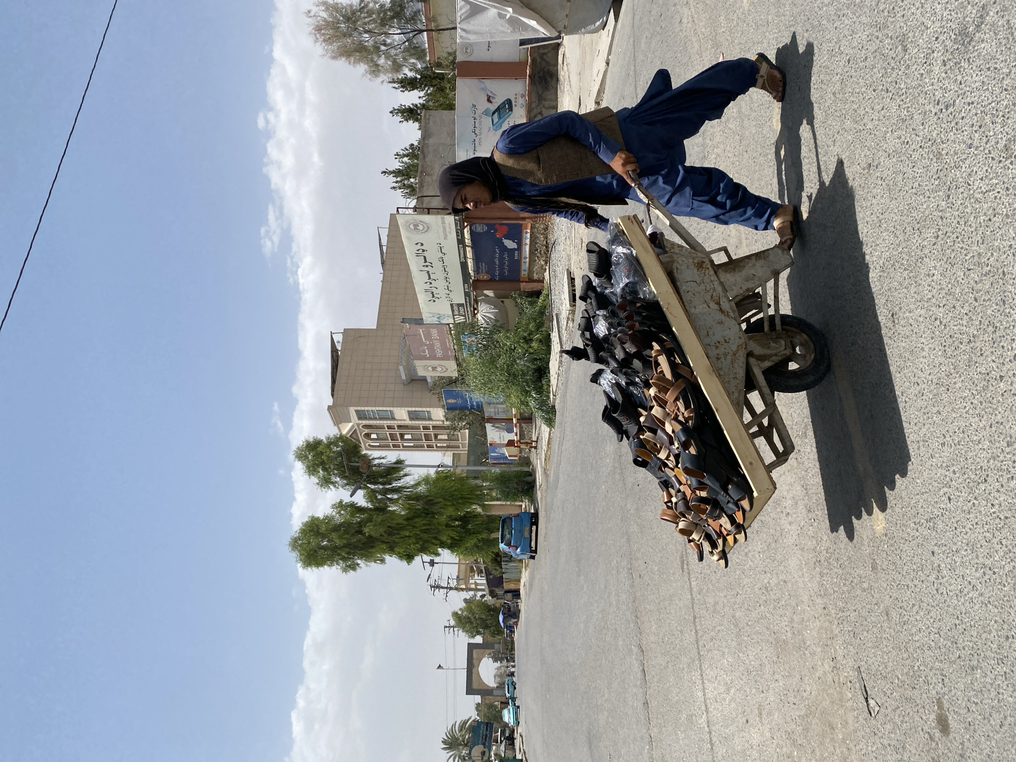 A man wearing a navy blue shalwar kameez and a dark vest pushes a wheelbarrow loaded with sandals down a paved street in Kandahar, Afghanistan. The wheelbarrow is old and appears rusted, with a wooden bed filled with various styles of sandals. The man is partially obscured by the wheelbarrow, but his legs and feet, clad in sandals, are visible as he walks. In the background, buildings line the street, including one with Arabic script signage. A blue car is parked on the side of the road. Trees and power lines are also visible. The sky is partly cloudy and bright, suggesting a sunny day. The overall scene depicts a commercial or transportation activity on a street in Kandahar.