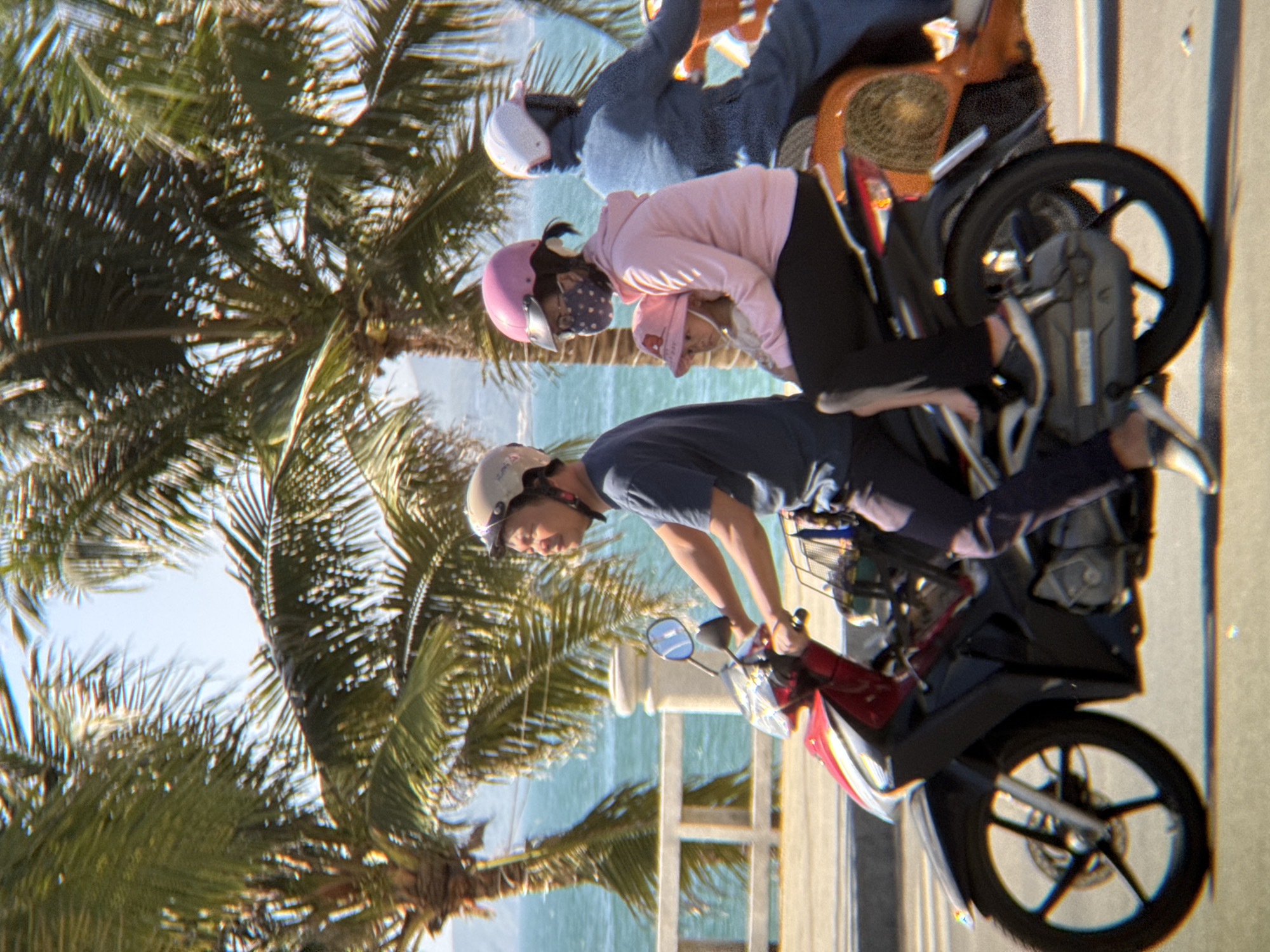 In Nha Trang, Vietnam, four people are riding on two motorcycles along a paved road by the sea, with palm trees in the background. The first motorcycle carries a man wearing a grey helmet and a dark t-shirt, driving. Behind him, on the second motorcycle, a woman in a pink helmet and a pink hoodie is holding a baby wearing a pink hat. To the right, another person is riding on the back of the second motorcycle, dressed in a dark blue garment and a white helmet. The motorcycles are moving forward. The sea is visible behind a white railing, and the sky is bright blue.