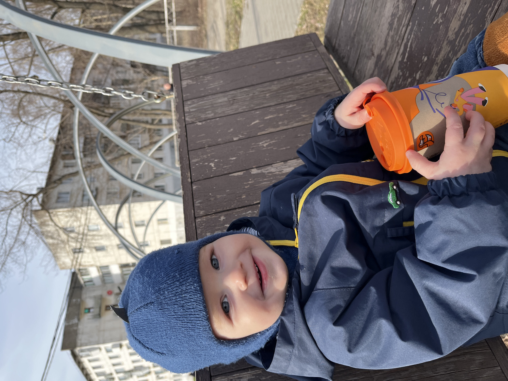 A young child, likely a toddler, wearing a blue knit hat and a blue jacket with a yellow stripe, is lying on a dark wooden bench. The child is smiling and holding an orange and yellow patterned cup with both hands. The scene appears to be outdoors, possibly in a playground or park. In the background, there's a metal structure with swirling tubes and a chain. Beyond that, there is a building with many windows and bare trees are also visible. This image was taken in Chaikovsky, Russia.