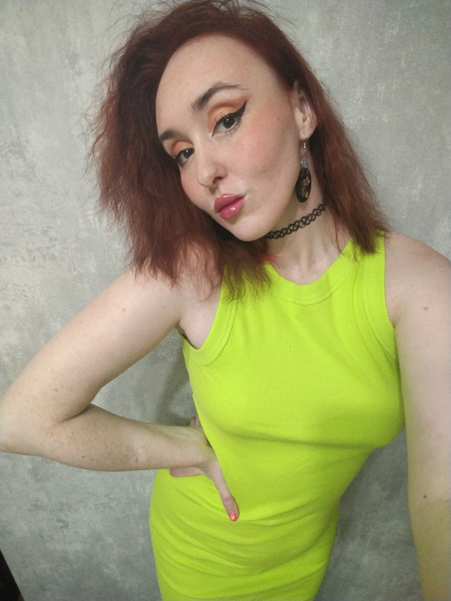 A woman with red hair, dressed in a neon yellow tank dress, poses in front of a gray wall. She is wearing black earrings and a choker. Her makeup includes winged eyeliner and glossy lips. Her left hand rests on her hip. The scene appears to be indoors, possibly in a residential setting. No location in Izhevsk, Russia, can be determined from the image.