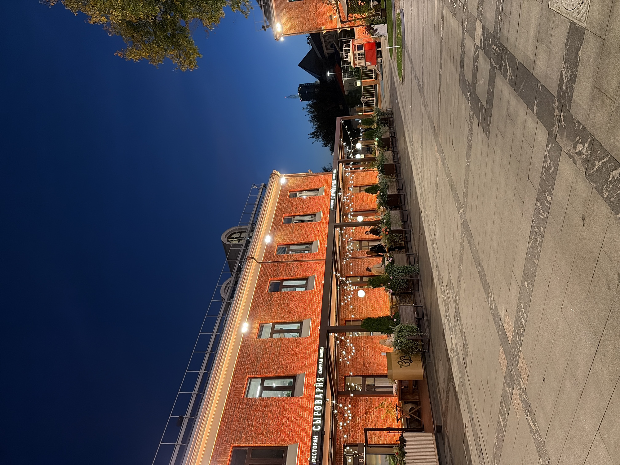 The image shows a brick building with an outdoor seating area in front of it, illuminated by string lights. The building has multiple windows and a red awning. Several people are seated at tables outside. The ground in front of the building is paved with large stone tiles. In the distance, there is another building and trees. A small red structure resembling a tram or trolley is visible to the right of the main building. The sky is a deep blue, indicating it is dusk or early evening. The scene appears to be in an urban setting. This location is the "Syrovarnya" restaurant on Bolshaya Nikitskaya Street in Moscow, Russia.