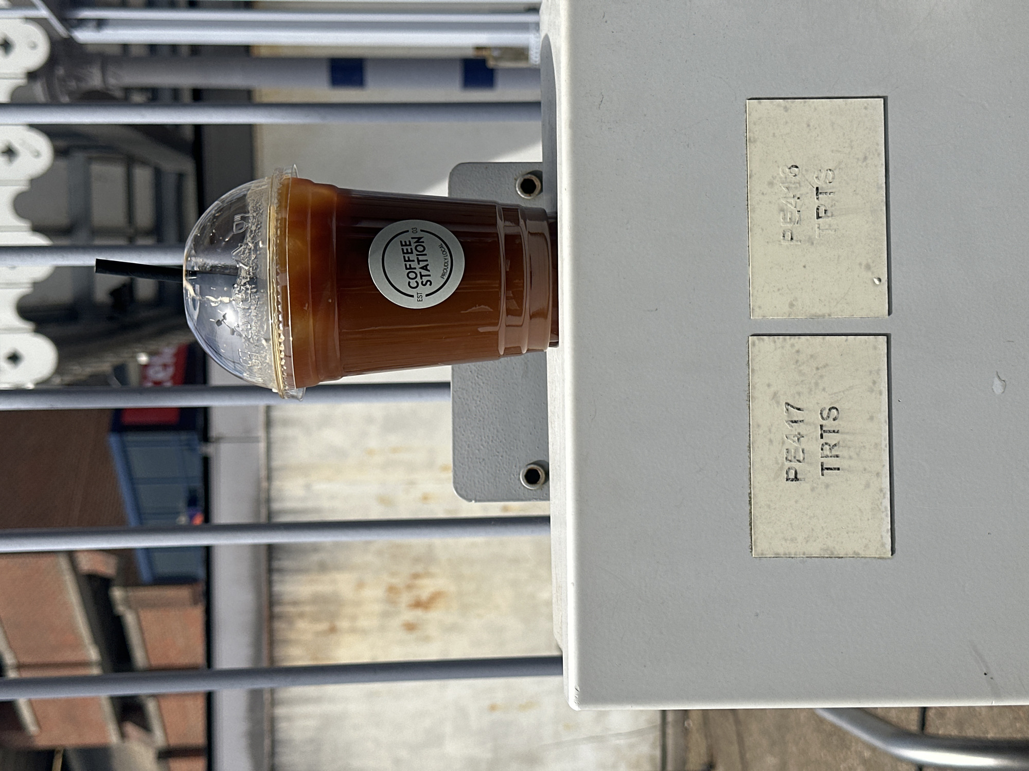 A cup of iced coffee from Coffee Station, a Tunbridge Wells, United Kingdom-based coffee shop, sits atop a gray electrical box. The cup has a clear plastic lid and a black straw. The label on the cup reads "Coffee Station" and "Proudly Local." The electrical box has two small, square labels that read "PE416 TRTS" and "PE417 TRTS." The background shows a gray metal structure and blurred buildings. The image is taken at an angle, with the electrical box in the foreground and the background slightly out of focus.