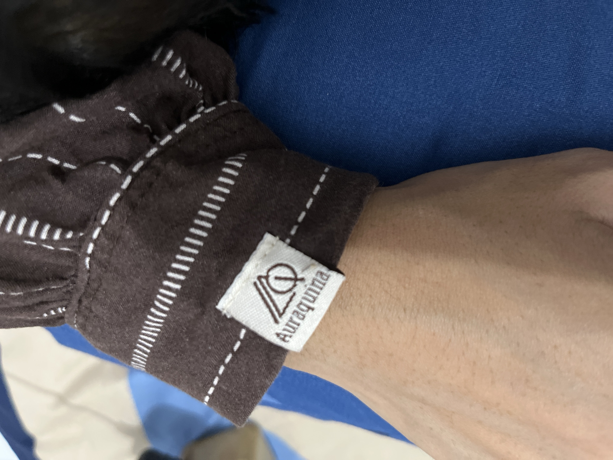 A close-up shot captures a person's light-skinned arm and wrist, adorned with a brown garment that features distinct white stitching. The fabric, possibly a sleeve or cuff, is decorated with patterns of dashed and solid parallel white lines. Attached to the brown garment, near the wrist, is a small, off-white rectangular clothing label. The label prominently displays a brown, stylized logo, which appears to be an intertwining of the letters "AQ" or "LO." Below the logo, the brand name "Auraquina" is vertically inscribed in brown lettering. A textured blue fabric fills the background, suggesting another piece of clothing or a soft surface. The lighting is bright and even, indicating a daytime setting, likely indoors. No specific activity is depicted beyond the static presentation of the clothing and arm. The image is taken in Lhokseumawe, Indonesia.