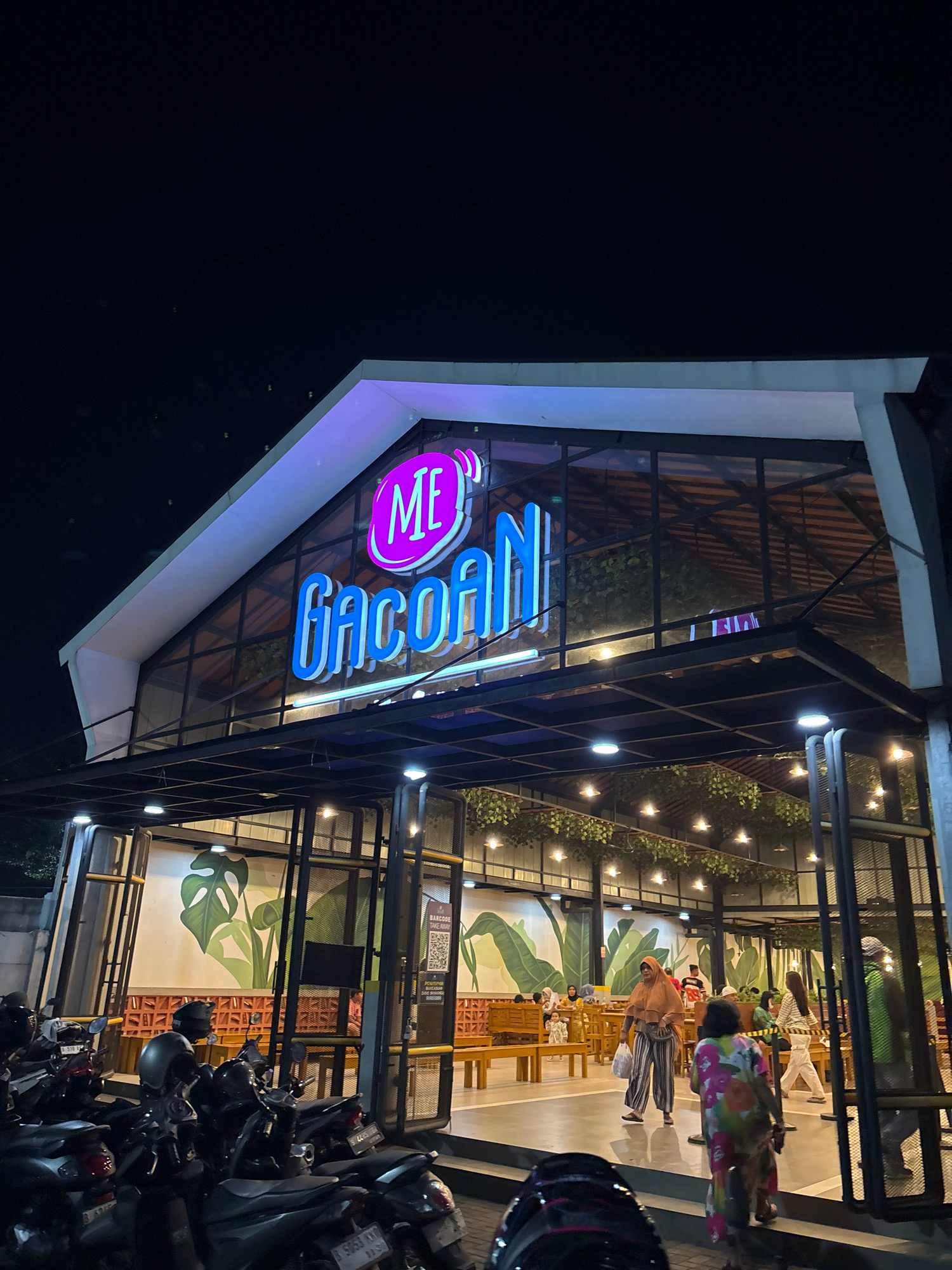 This is a photograph of the exterior of a restaurant named "ME GACOAN" in Bekasi, Indonesia, at night. The restaurant features a modern architectural design with a prominent glass facade and neon signage. The "ME GACOAN" logo is illuminated in bright blue neon, with a pink circular graphic above it. Inside the restaurant, diners can be seen sitting at wooden tables and benches. The interior is decorated with large wall paintings of tropical leaves and a hanging installation of artificial greenery with pendant lights.
In the foreground, several motorcycles are parked, suggesting that this is a popular mode of transportation for patrons. Some of these motorcycles are covered with helmets.
Several people are visible in the image. One woman, wearing a tan hijab and striped pants, is walking towards the right. Another person, wearing a colorful floral-patterned dress, is walking away from the camera. Other individuals are seated inside the restaurant. The overall atmosphere appears lively and inviting, despite being taken at night. The lighting from the restaurant's sign and interior creates a warm glow.