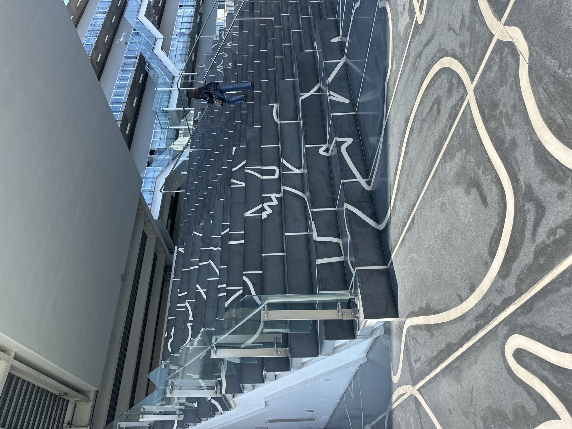 The image depicts a person walking on stairs with white graphic designs at the Soumaya Museum in Mexico City, Mexico. The museum's architectural elements, including glass railings and a distinctive exterior, are visible. The setting appears to be a bright, daytime scene.