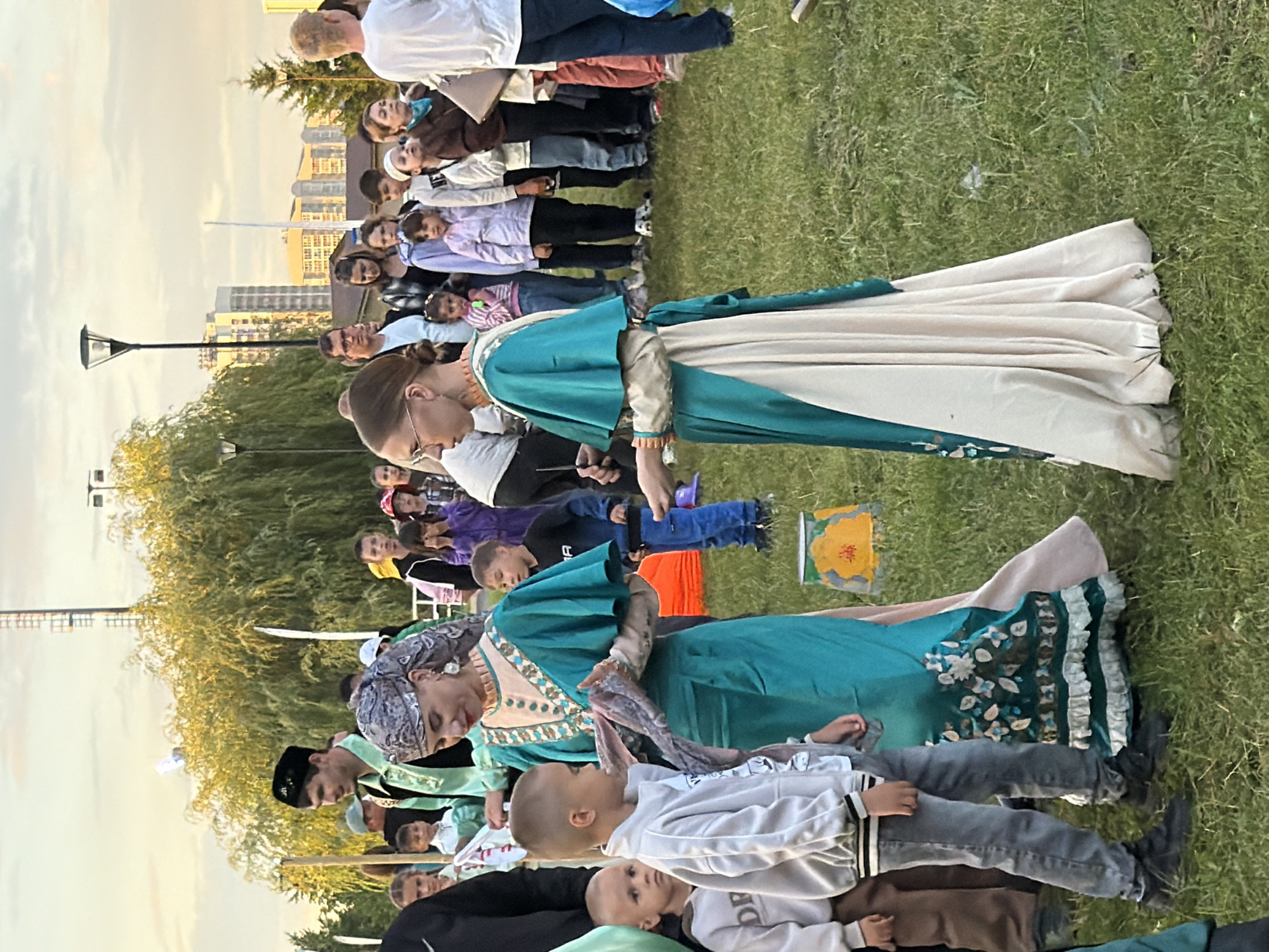 In an outdoor grassy area with trees in the background and city buildings visible on the horizon, a group of people are gathered. Two women in traditional, ornate dresses are interacting in the foreground. The woman on the left wears a teal and green gown with intricate floral embroidery and ruffles at the hem. She has a patterned headscarf and large earrings. The woman on the right wears a similar teal overdress with a cream-colored underdress. She has glasses and her hair is pulled back. A young boy in a grey hoodie and jeans stands to the left of the woman in the teal and green dress, facing her. Another young boy in a dark blue shirt and jeans stands further back on the right. Other individuals, including children and adults, are visible in the background, some standing and some in line. The lighting suggests it is either early morning or late afternoon.