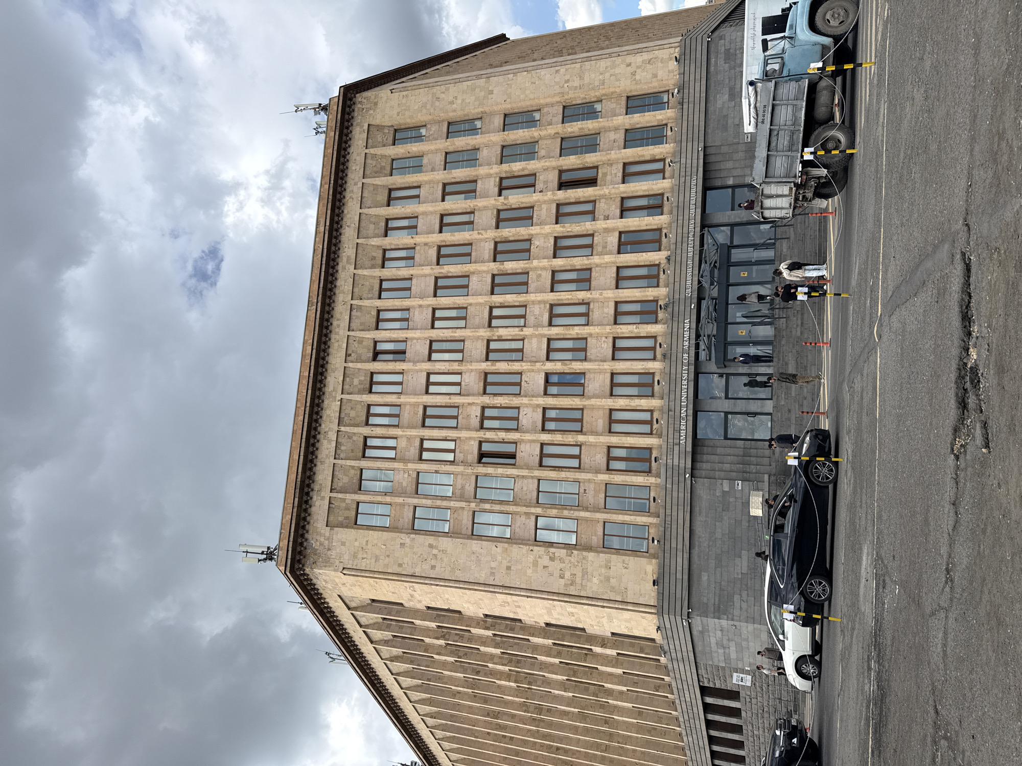 The image depicts the American University of Armenia building in Yerevan. It is a multi-story, rectangular structure with a light-colored facade and numerous windows. The building is adjacent to a street with parked cars and a truck near the entrance. People are present near the entrance, some of whom appear to be entering or exiting the building. The sky is overcast with cloudy conditions.