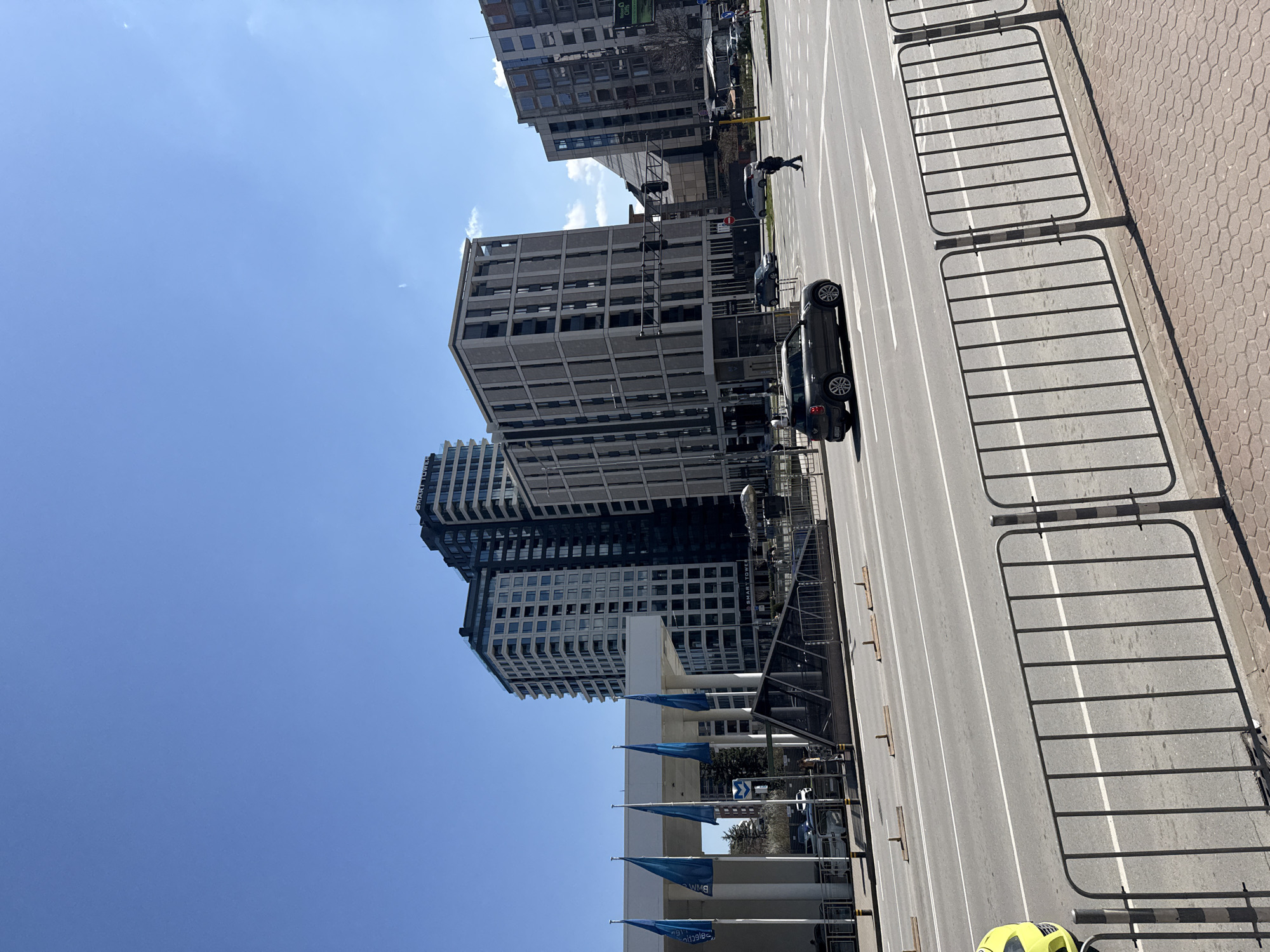 The image shows an elevated view of a street in Sofia, Bulgaria. Tall, modern buildings, some with blue flags at their base, line one side of the street. A dark-colored car is parked on the right side of the street, and a pedestrian is walking further down the road. Metal barriers are placed along the edge of the pavement on the right. The sky is blue with minimal clouds. The street has lane markings. The location is likely near business buildings in Sofia.