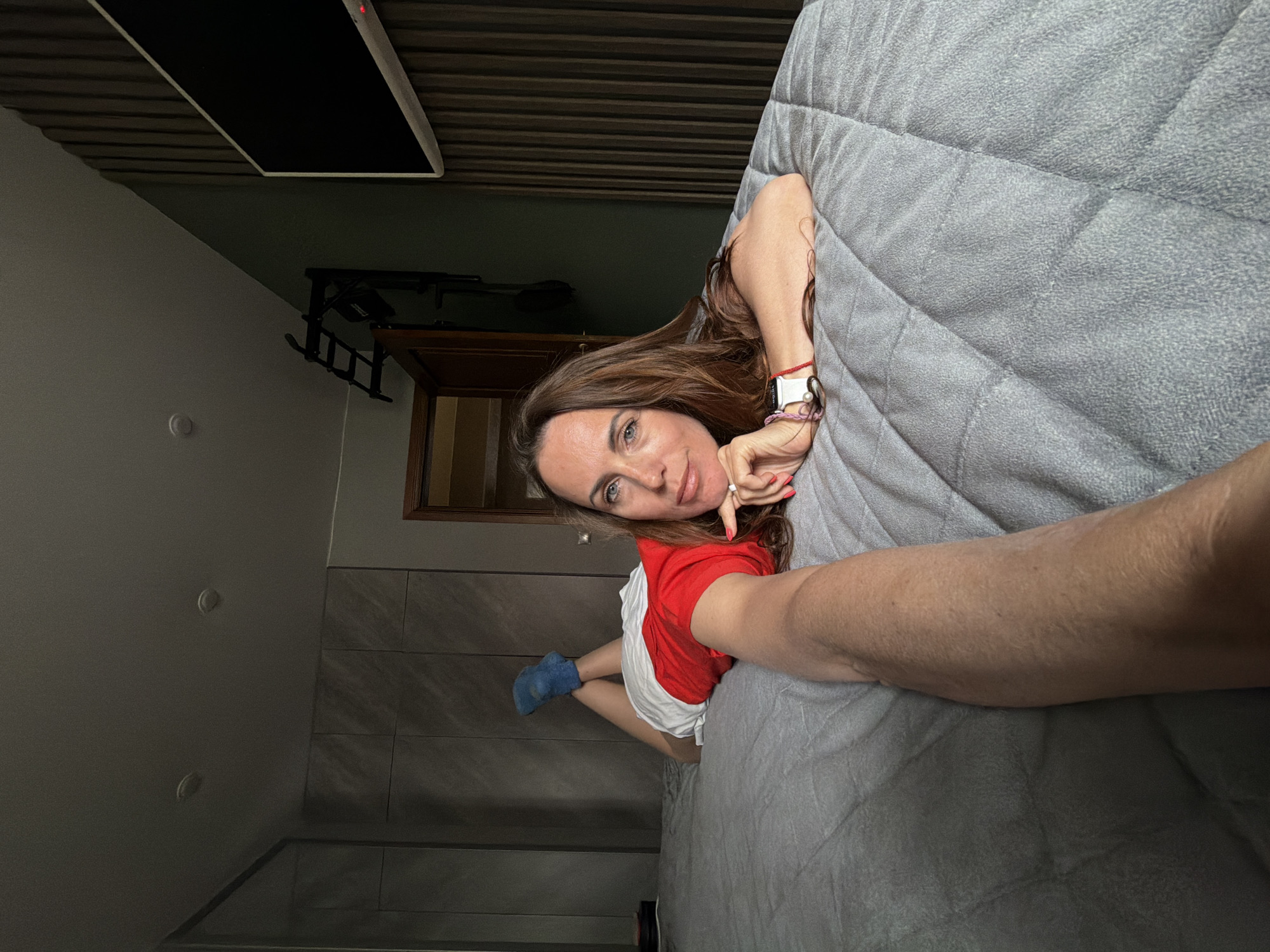A woman with long brown hair, wearing a red and white shirt and one blue sock, lies on a gray quilted bedspread. She is looking at the camera and touching her face with her right hand. A television is mounted on the wall, along with an exercise pull-up bar. A wooden framed door or opening is visible. The room has gray walls and a dark wood ceiling. There is no verifiable information to suggest this image was taken in Birobidzhan, Russia.
