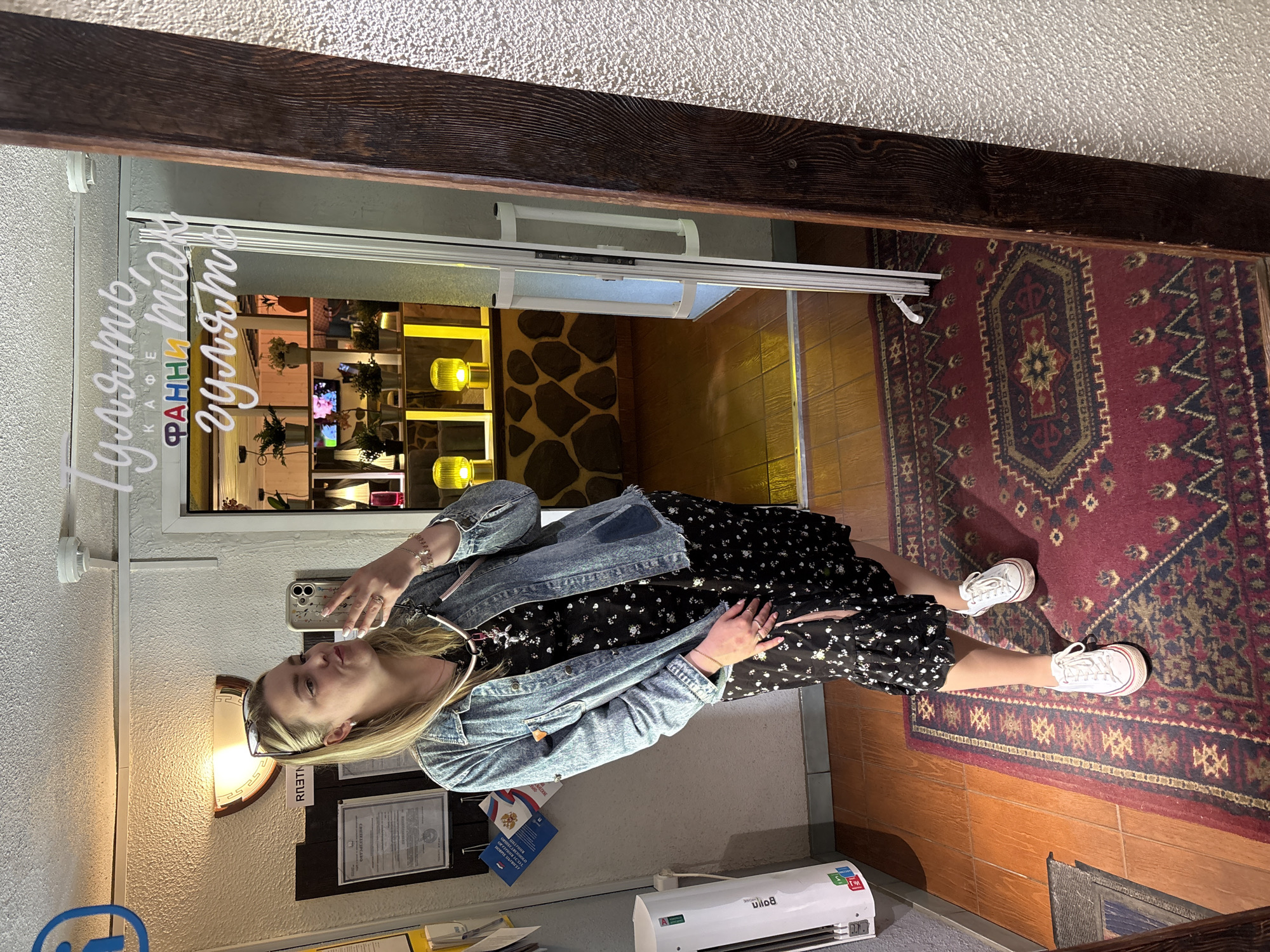 A woman stands in front of a glass door at the Fanny Mac Cafe in Chaikovsky, Russia. She is dressed in a denim jacket over a black floral dress, and white sneakers. She holds her phone to take a photo of herself in front of the restaurant. Behind her, inside the cafe, are decorative plants, illuminated lights, and a television. A patterned rug is visible on the floor. Outside the cafe, on the wall, are various documents and a small air conditioning unit.