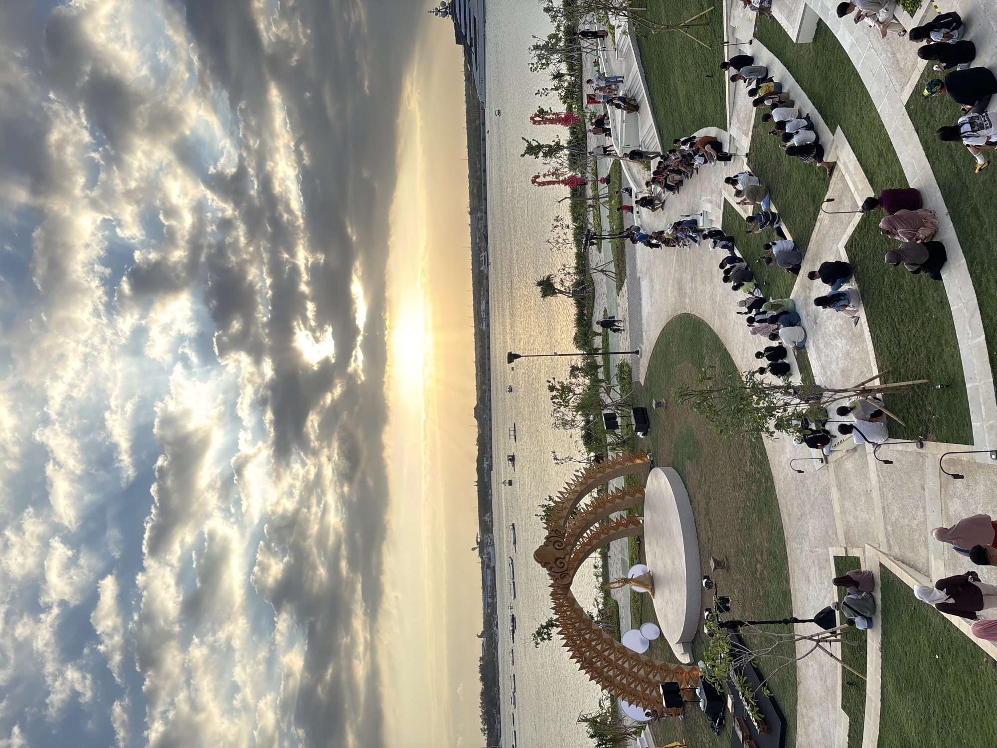 The image depicts an outdoor amphitheater setting by the water, likely during sunset. A stage is set up with a golden, arched structure resembling dragon scales. A single performer in a gold costume stands on the circular stage. The audience is seated on tiered, grassy steps overlooking the stage, facing the water. Several boats are visible on the calm body of water, and a large yacht is docked in the distance. The sky is filled with dramatic clouds, with the sun setting and casting rays of light through them. The scene appears to be from an event or performance in an urban coastal area of Indonesia.