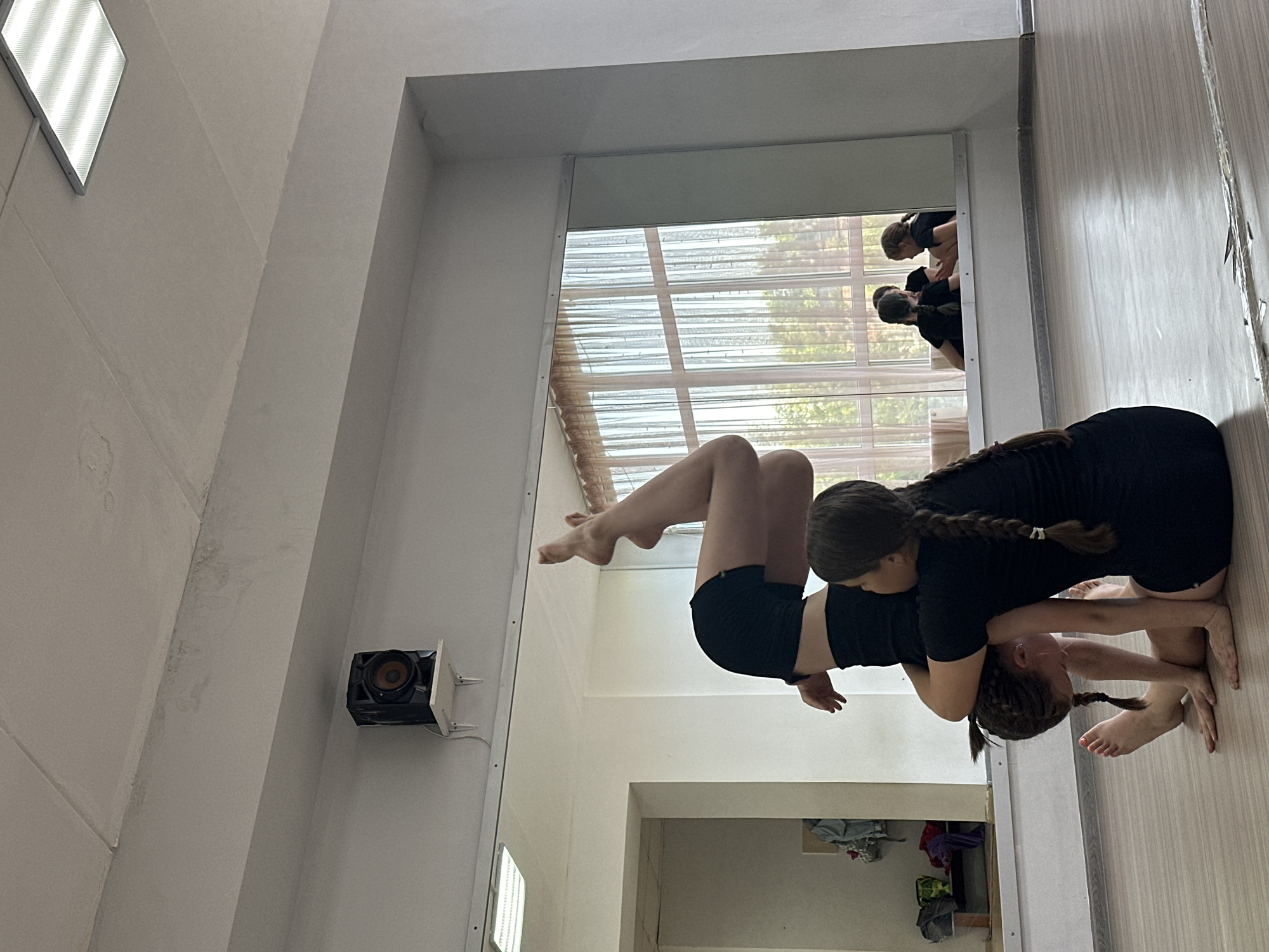 This image, captured in Votkinsk, Russia, shows two young girls engaged in a flexible acrobatic or dance pose within what appears to be a studio. Both girls have braided hair and are dressed in black athletic tops and shorts. One girl is leaning back, supported by the other, with their bodies intertwined in a balanced posture on a light-colored floor.
A large mirror covers a significant portion of the wall, reflecting the practice space. In the mirror, at least two other individuals, also in black attire, are visible, suggesting a group training session. The reflection also reveals window blinds and outdoor greenery, indicating daytime.
The studio has plain light-colored walls and ceiling. A rectangular light fixture is mounted on the ceiling, and a black speaker is attached to the wall on the left. Some personal items or clothing are visible on the floor in the foreground. The overall scene suggests a dedicated and focused training environment. No visible text is present in the image.