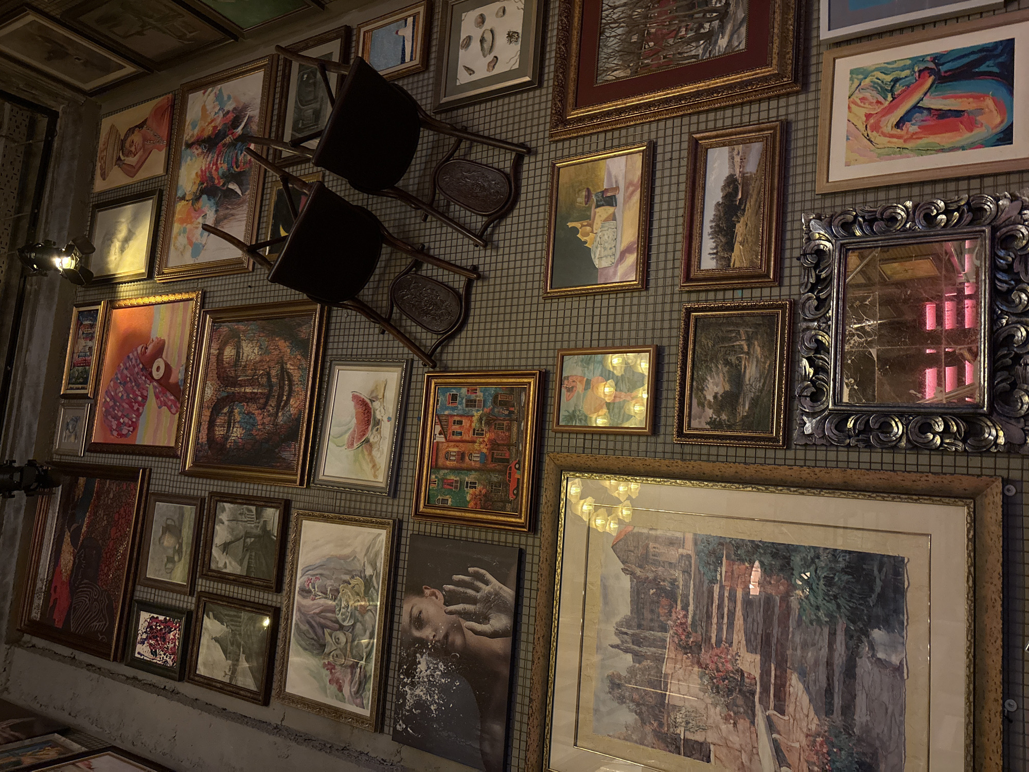An interior wall, likely in Kazan, Russia, is densely covered with framed artwork of various styles, sizes, and subjects. Two chairs are mounted on the wall among the frames. The wall surface appears to be a grid-like metal or wire mesh.
