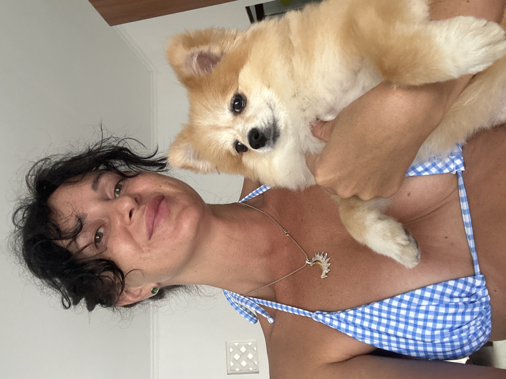 A woman with dark, curly hair and light brown eyes, wearing a blue and white checkered bikini top and a silver sun pendant necklace, is holding a light brown Pomeranian dog. The woman is smiling and looking at the camera. The dog is looking to the left of the frame. The background is a plain white wall. The image appears to have been taken indoors, likely in a residential setting in Kuta Selatan, Indonesia.