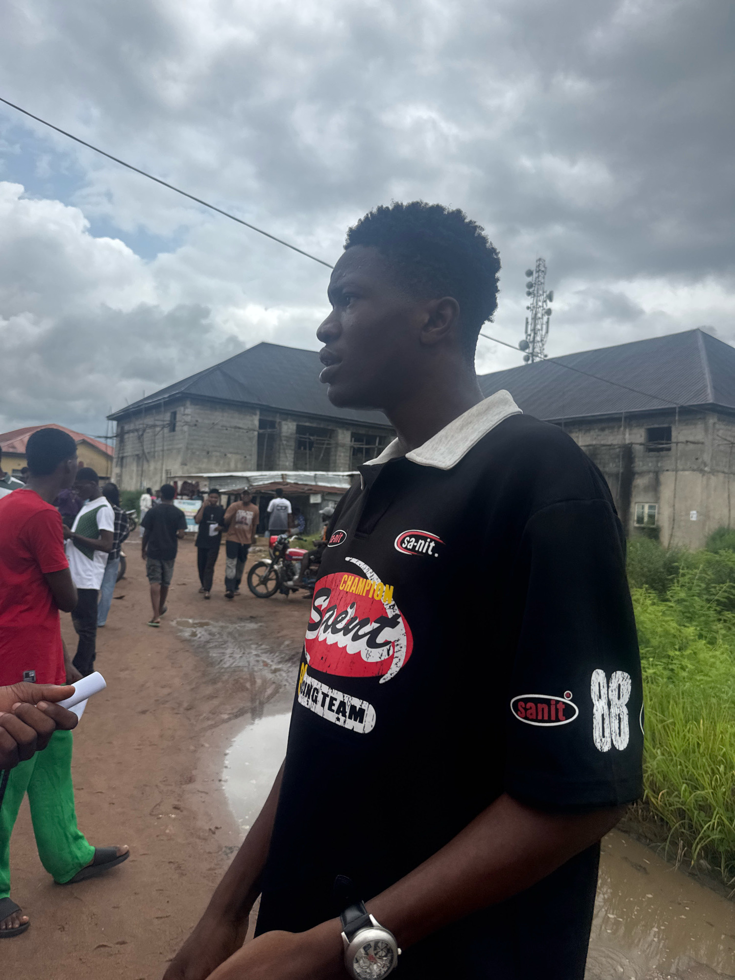 In Ihiagwa, Nigeria, on a cloudy day, a young Black man is captured in a close-up profile shot, looking to his right. He wears a black polo shirt with a white collar, emblazoned with a red and white logo on the left chest that reads "san-it.". Below this logo, a larger graphic features stylized red text that appears to say "Champion" with a white outline, and "Speed" in white text. Towards the bottom of the graphic, "KING TEAM" is visible in white text. On the right sleeve, the number "88" is printed in white, accompanied by another "san-it." logo.
The man is standing on a dirt road, which is partially covered in puddles, indicating recent rain. In the background, several other individuals are seen walking away from the camera. To the left, a person in a red t-shirt and green pants is visible, holding a white rolled-up object. Further back, there are two-story buildings under construction, one with exposed concrete and rebar, and another with a dark roof. A telecommunications tower stands tall against the overcast sky. Green vegetation borders the right side of the frame. A red motorcycle is parked near a building in the mid-ground. The overall scene depicts a somewhat unpaved, possibly rural or semi-urban environment.
