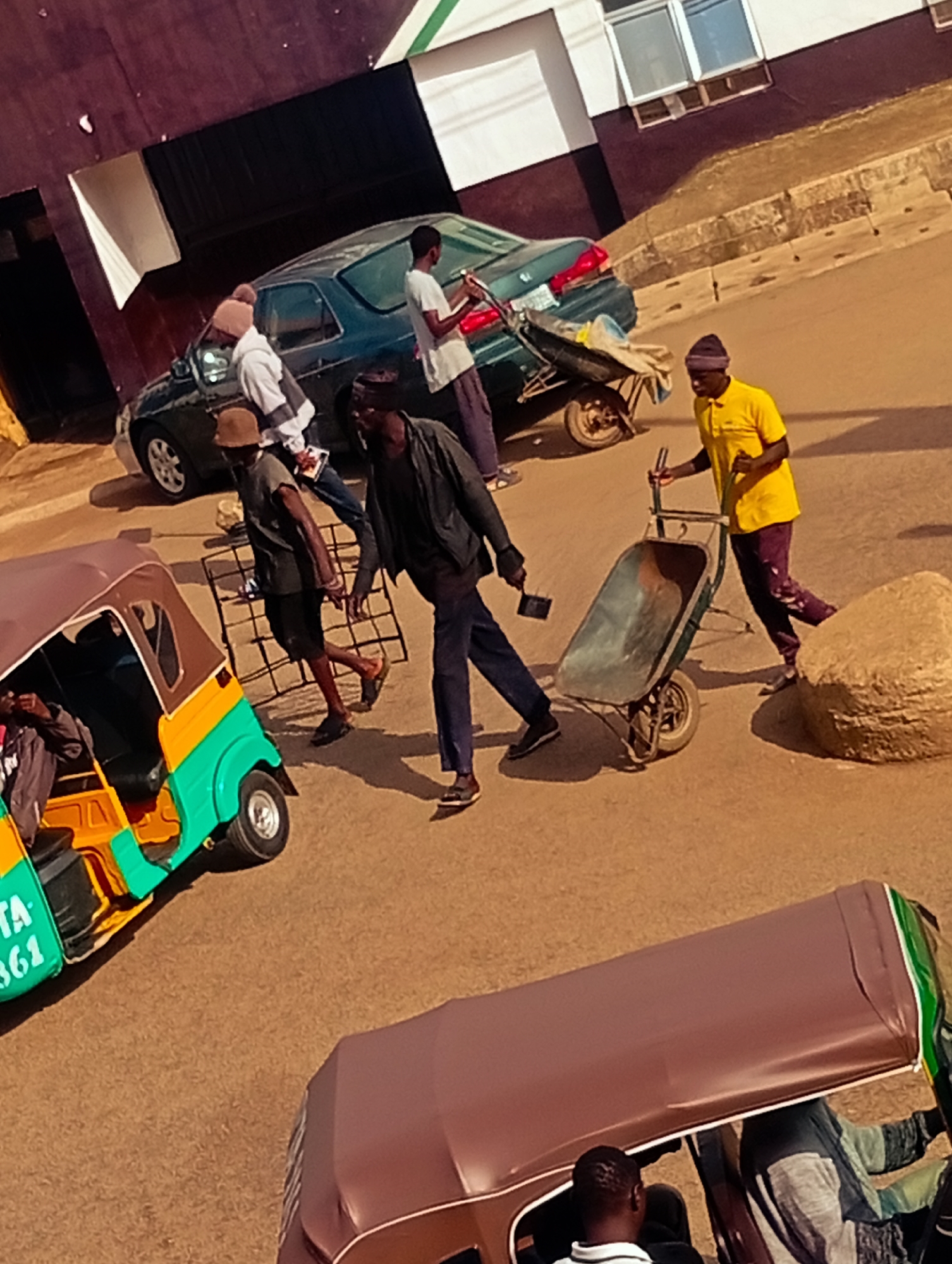 This is a street scene in Bukuru, Nigeria, with several people and vehicles. In the foreground, a three-wheeled auto-rickshaw, brightly colored in green and yellow, is parked on the right side of the frame, with a driver visible inside. Further back, a dark-colored sedan is parked. Two men are interacting with a wheelbarrow. One man, wearing a yellow shirt and dark pants, appears to be pushing the wheelbarrow, while another man in a dark jacket and blue pants walks alongside, holding a shovel. Two other individuals are near the sedan, one wearing a white hooded jacket and a beanie, and another in a brown hat. The environment appears to be an urban street with a building in the background that has dark and white sections, and windows. The ground is a sandy-brown color, typical of a dusty road.