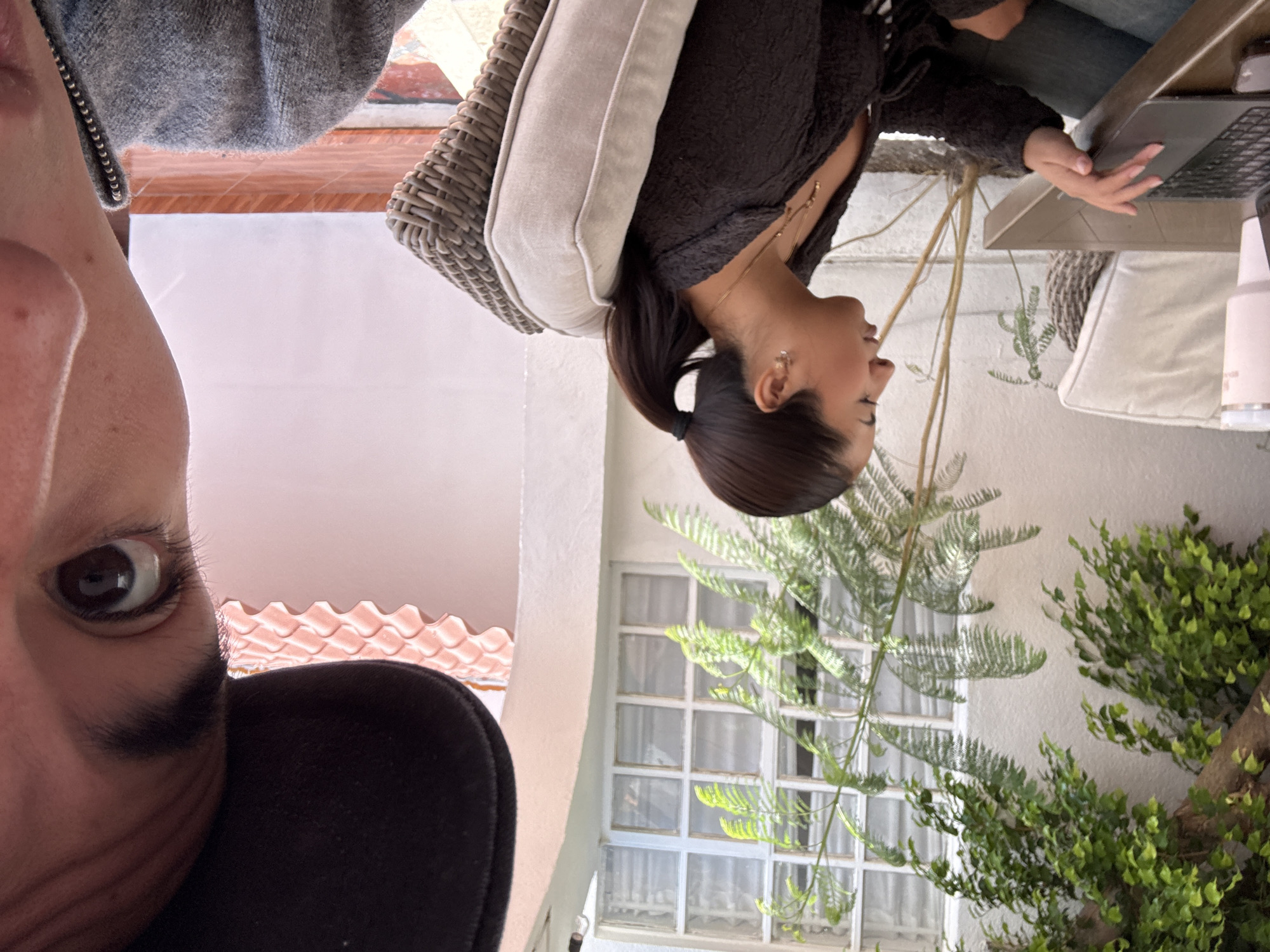 The image captures two people in an outdoor setting, likely a patio or garden area. One individual, positioned in the lower left corner, is captured in a close-up selfie, focusing on their eye and part of their face. They are wearing a black garment or accessory, potentially a hat or jacket. The other person is seated in a wicker chair with a light-colored cushion, facing away from the camera. They are wearing a dark top, gold necklaces, and have their dark hair pulled back in a ponytail. They appear to be working on a laptop, with their hands positioned on the keyboard. A white water bottle stands on the table next to the laptop.
The background includes white walls, windows with white frames and curtains, and lush green foliage, including a tree and various plants. The presence of red roof tiles above the white wall suggests a traditional architectural style, characteristic of Puebla, Mexico.