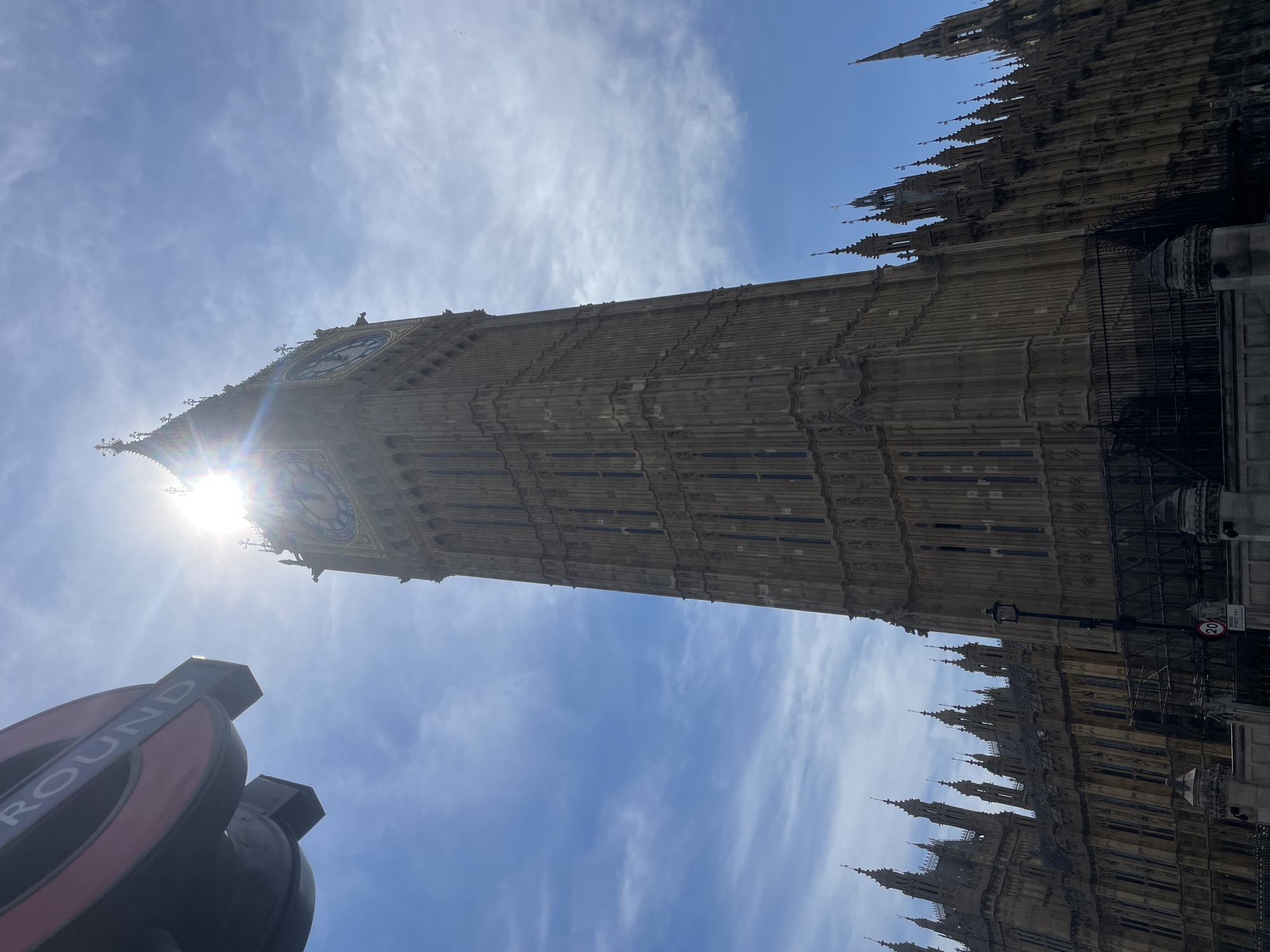A wide-angle, low-angle shot captures the Elizabeth Tower, commonly known as Big Ben, in London, United Kingdom. The iconic clock tower, part of the Palace of Westminster, dominates the frame, its stone facade detailed with Gothic architecture. Sunlight streams through the clouds, creating a lens flare effect near the top of the tower, partially obscuring the clock faces. In the foreground, a portion of a London Underground sign, with the word "ROUND" visible, juts in from the upper left corner. The sky is a vibrant blue, with scattered white clouds. The surrounding buildings of the Palace of Westminster are visible to the left and right of the tower, also displaying intricate architectural details and spires. A black metal fence runs along the base of the buildings. A street lamp and a circular speed limit sign (20) are also present in the lower portion of the image. No people are visible.