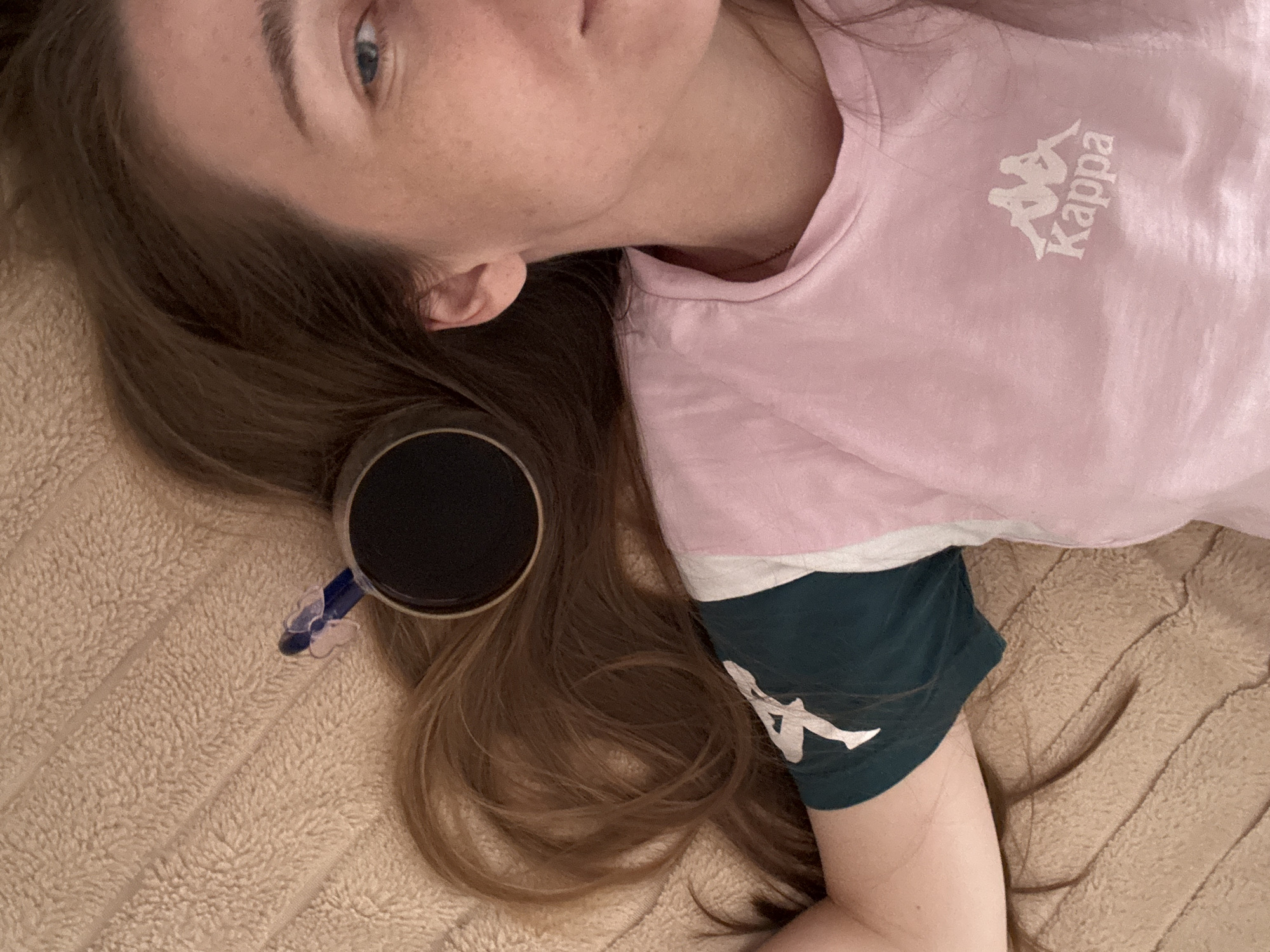 A person with brown hair is lying on a beige blanket with their head resting on their hair. They are wearing a pink t-shirt with a white Kappa logo on the right side of the chest and a dark green t-shirt underneath with a white logo on the left sleeve. A black cup of coffee is placed on the person's hair, with a blue hair clip attached to the cup's rim. The person's face is partially visible on the right side of the image, showing their right eye, eyebrow, and cheek with freckles. The background is a soft, textured beige blanket.