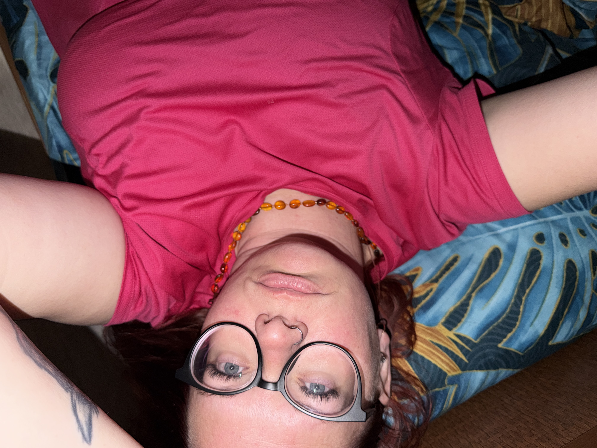 A woman is lying on a patterned blue and gold pillow or cushion. She is wearing a red shirt and a necklace of amber-colored beads. She is also wearing black-framed glasses. The woman is indoors. The location in the city of Chaikovsky, Russia is not evident.