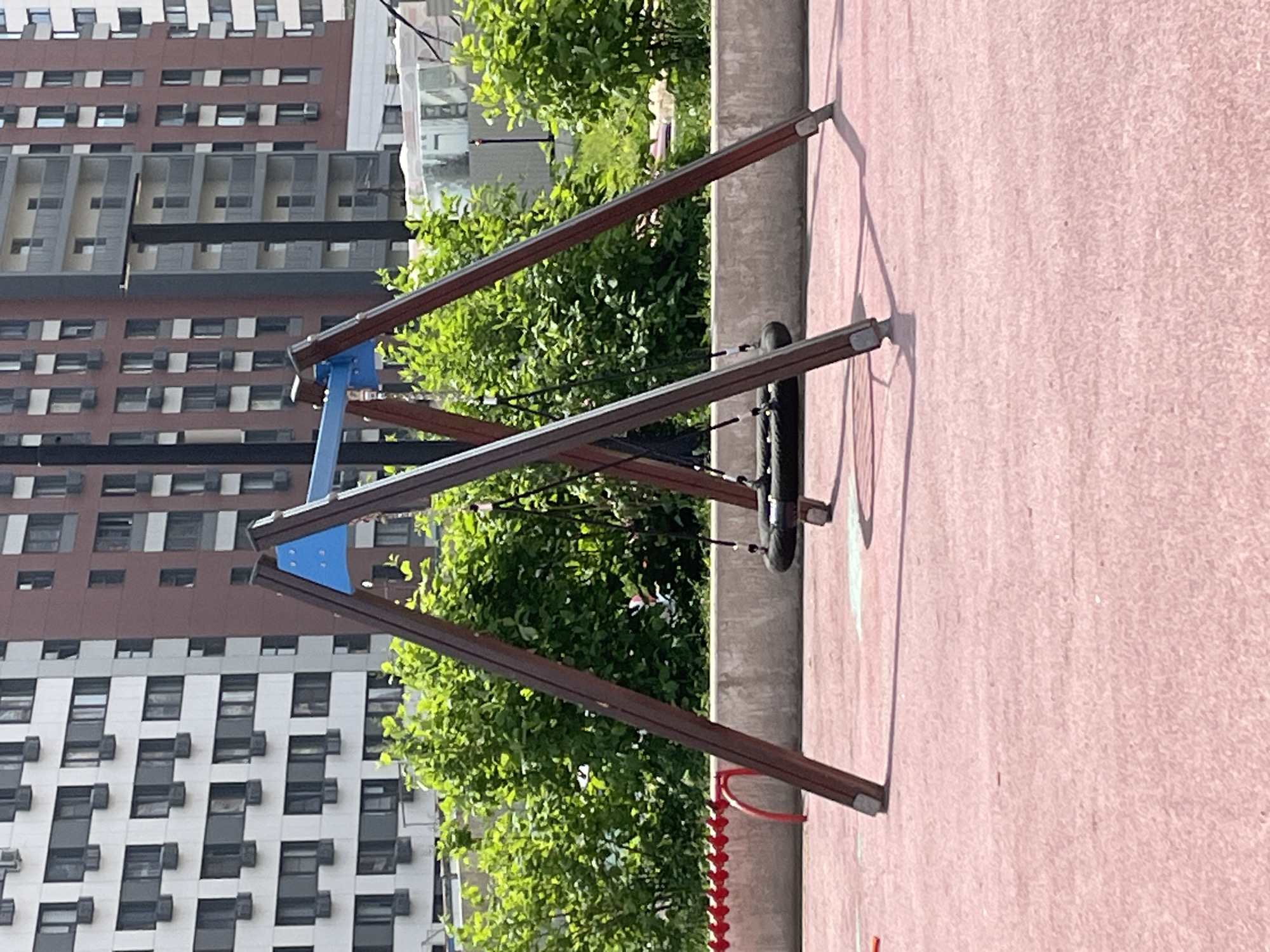 A landscape-oriented, medium-shot photograph depicts a tire swing at a playground, likely in Moscow, Russia, though this cannot be definitively determined from the image. The swing set is constructed with brown wooden supports and blue connecting pieces. The tire itself is black and suspended by ropes. The swing set is set against a backdrop of lush green foliage and a tall, modern apartment building with many windows. The ground beneath the swing set is a light pink or reddish surface. A red coiled object is also visible next to one of the supporting posts. There are no people visible in the image.