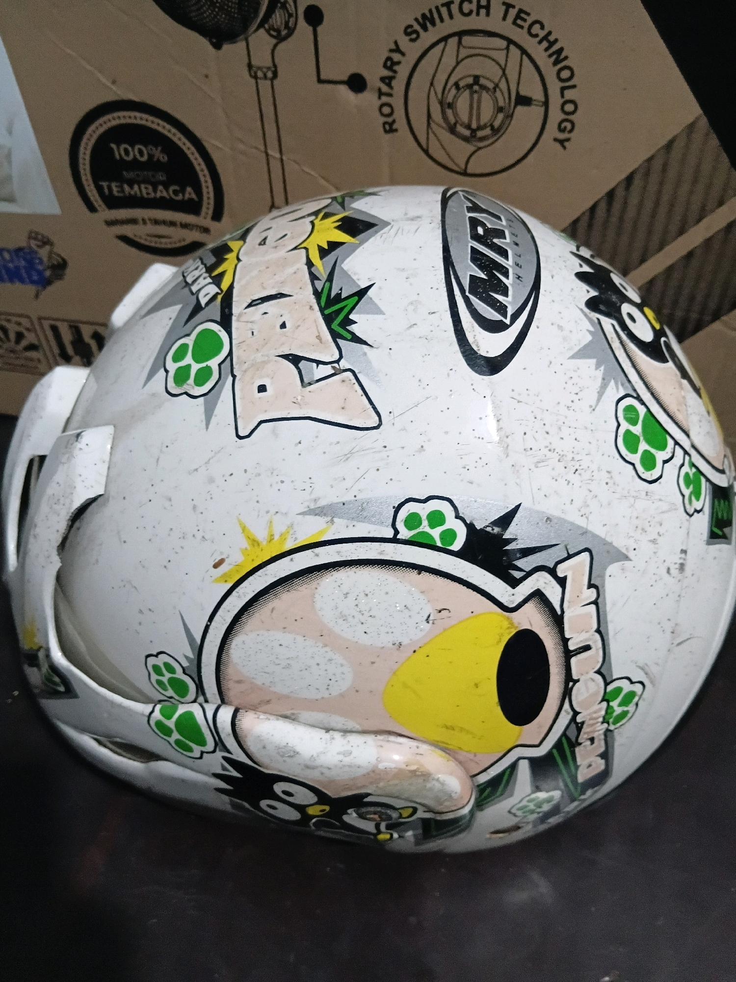 This is a close-up, eye-level, indoor photograph of a white, highly-decorated motorcycle helmet, likely a full-face helmet, resting on a dark, flat surface. The helmet is positioned at an angle, facing the viewer's right, with the top and right side prominently displayed. The helmet is covered in various cartoonish stickers and graphics.
Prominent graphics include:
- A large, light-colored sticker with black outlines depicting a character's face with large, rounded eyes and an open mouth. This graphic extends across a significant portion of the helmet's side.
- A white sticker with black and silver lettering that reads "MRY HELMETS."
- Scattered green paw-print graphics.
- Other black and white cartoon characters and abstract shapes.
- The helmet itself appears worn and scratched, with numerous small specks and scuffs visible on its white surface. There is also a notable crack or damage on the left side of the helmet, near the opening for the face shield.
In the background, out of focus, are parts of a cardboard box or packaging. One visible section of the box has text and graphics related to "ROTARY SWITCH TECHNOLOGY" and a circular diagram. Another section of the box displays a circular emblem with text that reads "100% MOTOR TEMBAGA" and "GARANSI & TAHUN MOTOR." These visual elements suggest the helmet may be new or recently purchased and that the photograph was taken in a retail or storage setting in Cikal, Indonesia. The lighting in the scene is dim, casting shadows that highlight the texture and details of the helmet.