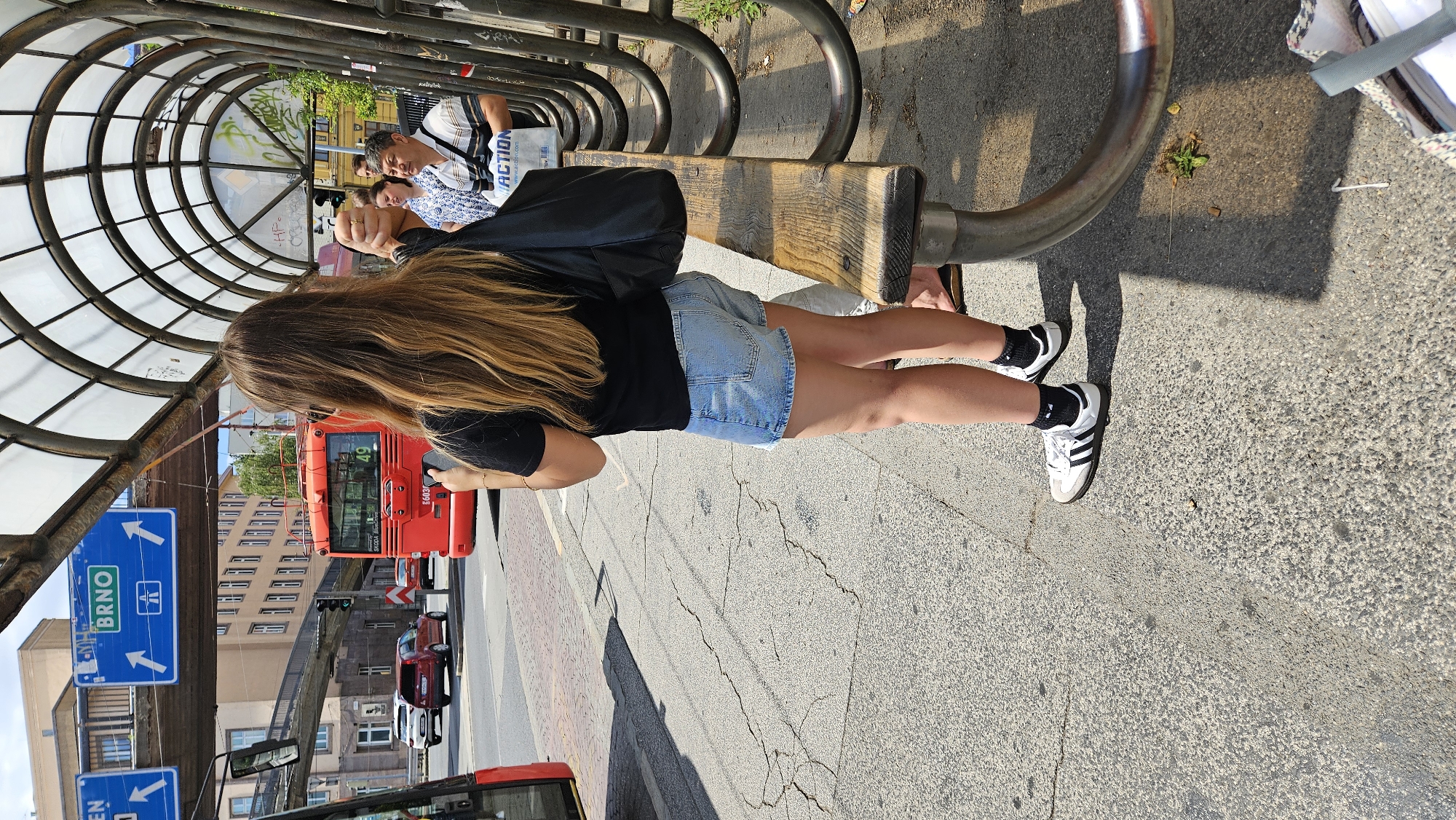 A rear view of a young woman with long, blonde hair, wearing a black t-shirt, denim shorts, black and white Adidas sneakers, and black socks, stands at a bus stop. She is holding a smartphone in her right hand and a black bag in her left hand. To her left, a red bus with the number "49" displayed on its front is visible. In the background, there are signs indicating directions to "BRNO" and other locations, as well as buildings and a pedestrian bridge. A man and a woman are also visible in the background, standing near the bus stop. The bus stop features a curved, transparent roof supported by metal structures and a wooden bench. The ground is paved with asphalt, and there are cracks in the pavement. This scene is at a bus stop in Bratislava, Slovakia.