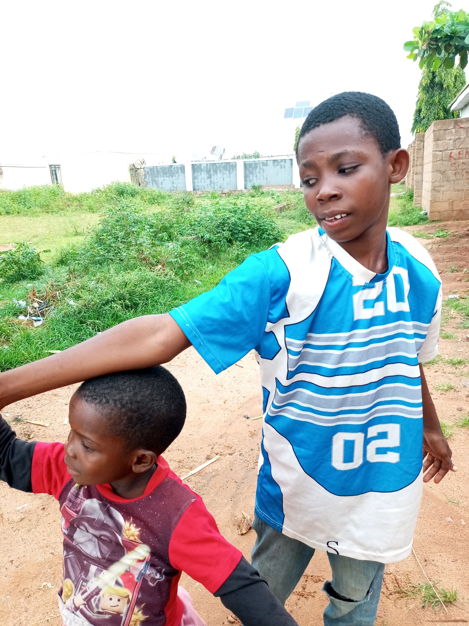 In Tempure, Nigeria, two young boys are the subjects of this medium-shot photograph taken outdoors. The boy on the right, older and taller, is wearing a blue and white t-shirt with the numbers "20" and "02" visible. His arm is resting on the head of the younger boy, who is on the left and is wearing a red t-shirt with a graphic print. The background consists of dry, sandy ground, sparse green vegetation, and a low, light-colored wall. The sky is overcast.