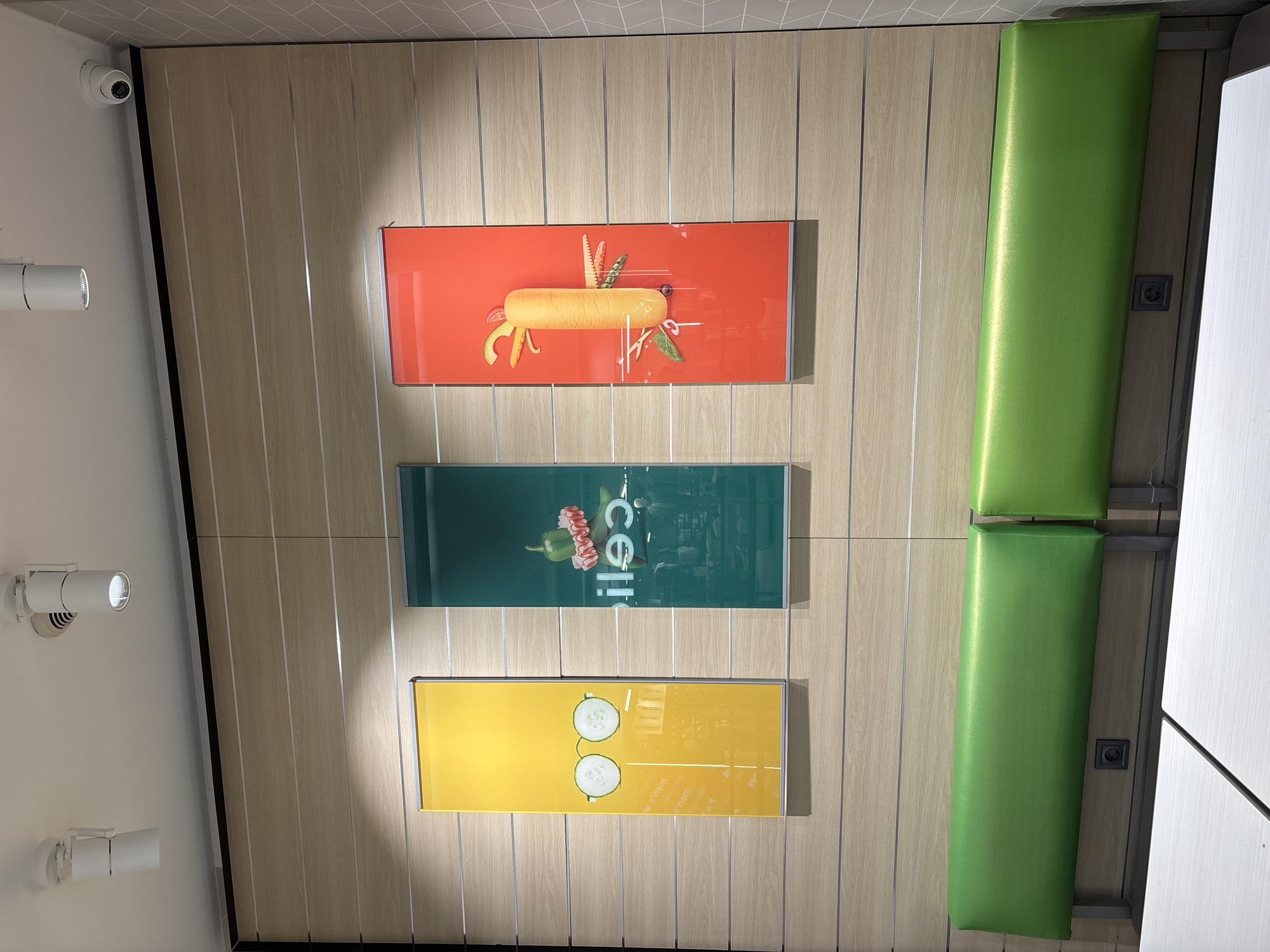 The image shows an interior wall with three horizontal rectangular pictures aligned vertically on a light-colored wooden paneled wall. The top picture is orange and features an image of a sandwich. The middle picture is green and the word "Celli" is visible. The bottom picture is yellow and depicts cucumber slices resembling eyeglasses. To the right of the pictures are two vertical green padded cushions or backrests. The top left of the image contains a security camera and spotlights. The image was taken in a fast food restaurant. The location is likely in Sofia, Bulgaria.