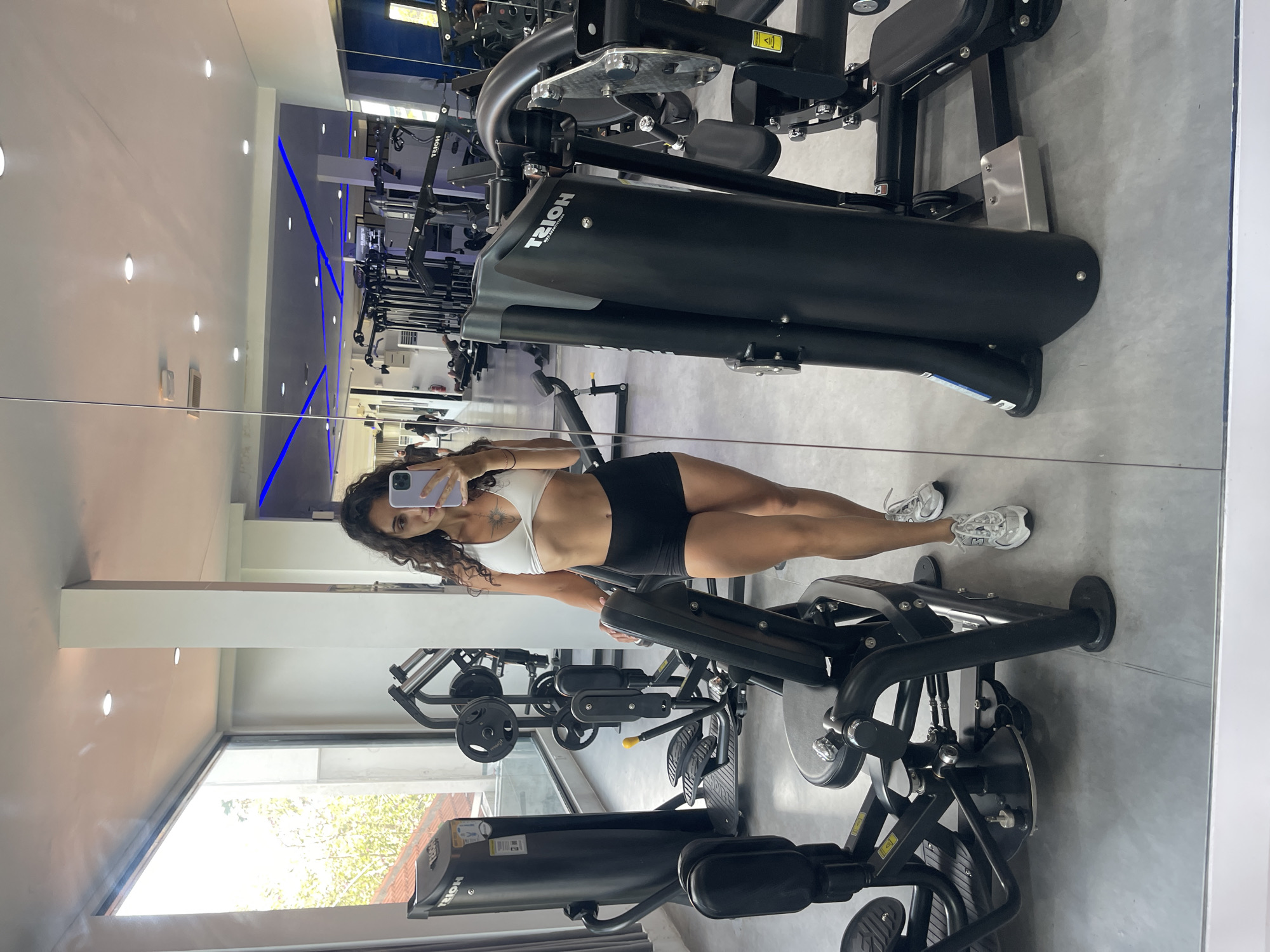 A young woman with curly dark hair and a navel tattoo is taking a mirror selfie in a well-lit gym. She is wearing a white sports bra and black athletic shorts, with white sneakers. The gym features modern black and gray weight machines, with a large window to the left offering a view of trees and sunlight. Blue neon lighting accents the ceiling in the background. The floor is a light gray, and the walls are white. The woman is positioned in the center foreground, holding a purple phone to capture her reflection. Several pieces of gym equipment, including weight machines and weight plates, are visible around her. Reflections in the mirror show other gym-goers and more equipment.