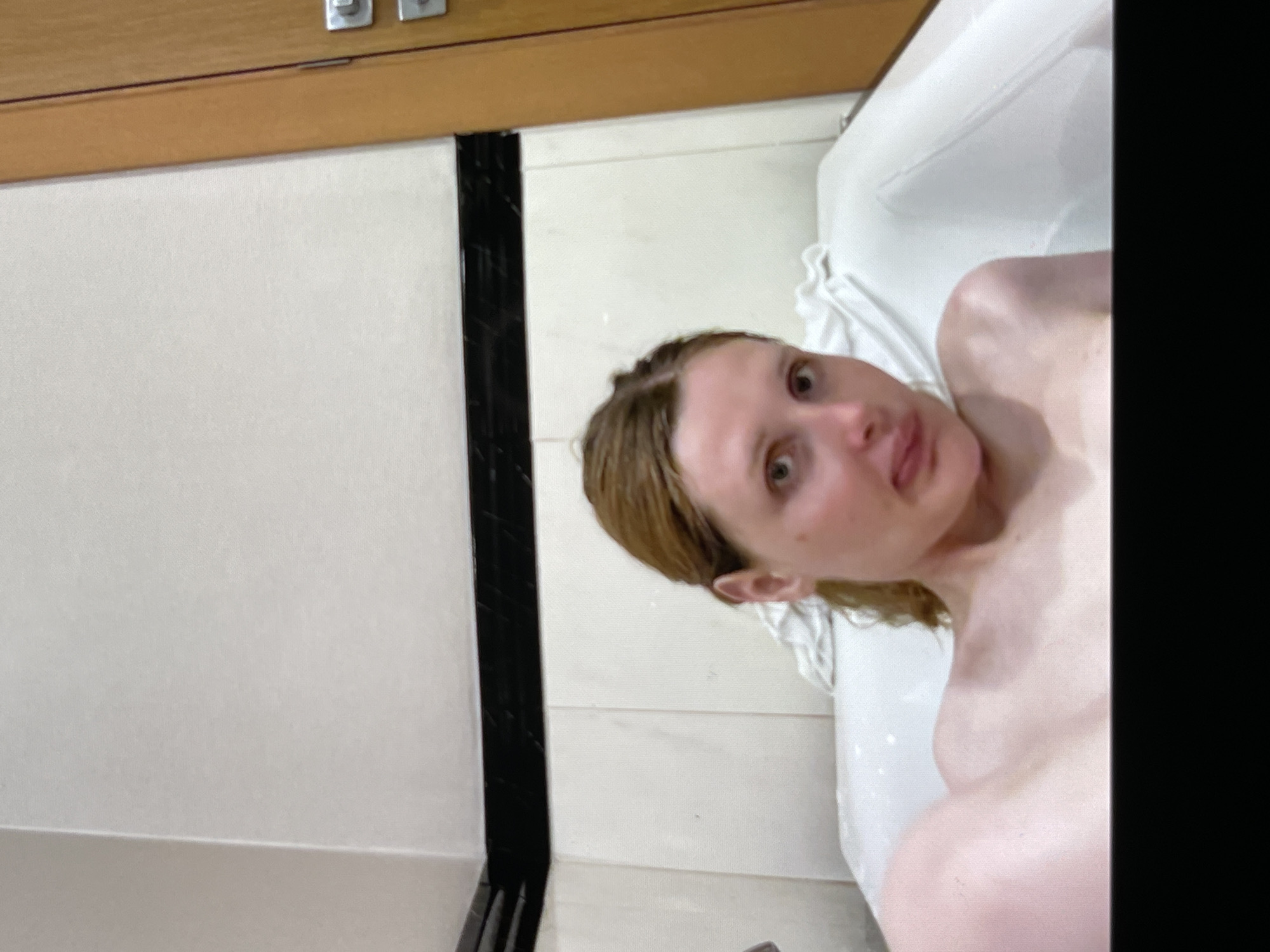 A woman with shoulder-length blonde hair is taking a selfie in a bathtub. Her head is tilted to the left and her mouth is slightly open, with her tongue sticking out. She is wearing a white towel around her shoulders. The bathtub is white and appears to be filled with water. The background is a tiled wall with a black border. There is a wooden door with a doorknob on the right side of the image. The lighting is soft and natural.