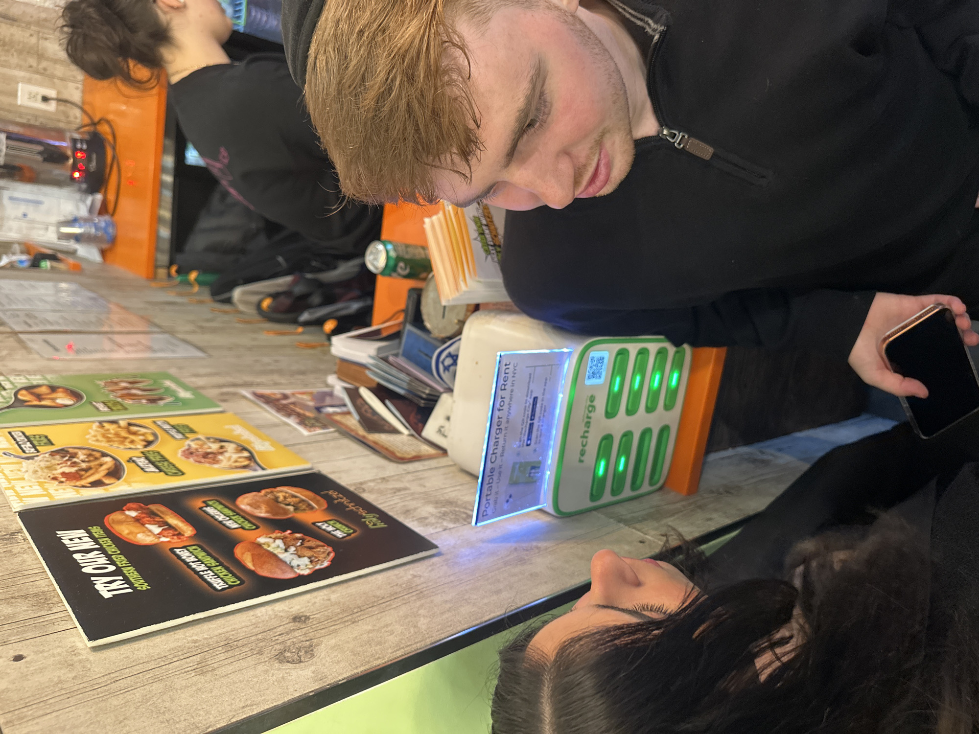 The scene depicts the interior of a Holy Schnitzel fast-casual restaurant located in New York City, United States of America. In the foreground, two individuals are present. On the right, a male with reddish-brown hair and a black quarter-zip shirt holds a gold-framed smartphone, screen-down, in his left hand, looking towards the left. On the left, partially cropped, a female with dark hair and a dark top faces right, looking towards the man or the objects between them. In the background right, a third individual, a female with dark hair in a bun and wearing a black shirt with a pink emblem, is seen from behind, attending to items on an orange counter.
The establishment features light wood-paneled walls, on which multiple laminated menu boards are displayed. One prominent black menu advertises "TRY OUR NEW SOUTHERN FRIED CHICKEN ITEMS," including "TRUFFLE HOT HONEY CHICKEN SANDWICH" and "NEW ORLEANS CHICKEN SANDWICH (The Holy Crunch)," bearing the "@holyschnitzel" brand. Other yellow and green menu boards show items such as "BUFFALO CHICKEN FRIES." Below these menus, mounted on an orange shelf, is a white "recharge" portable charger rental station, illuminated with blue and green indicator lights, displaying "Portable Charger for Rent" and "Return it anywhere in NYC." On the orange counter, a green beverage can, stacks of paper materials, and a silver countertop appliance with red indicator lights are visible. A white electrical outlet is mounted on the wall.