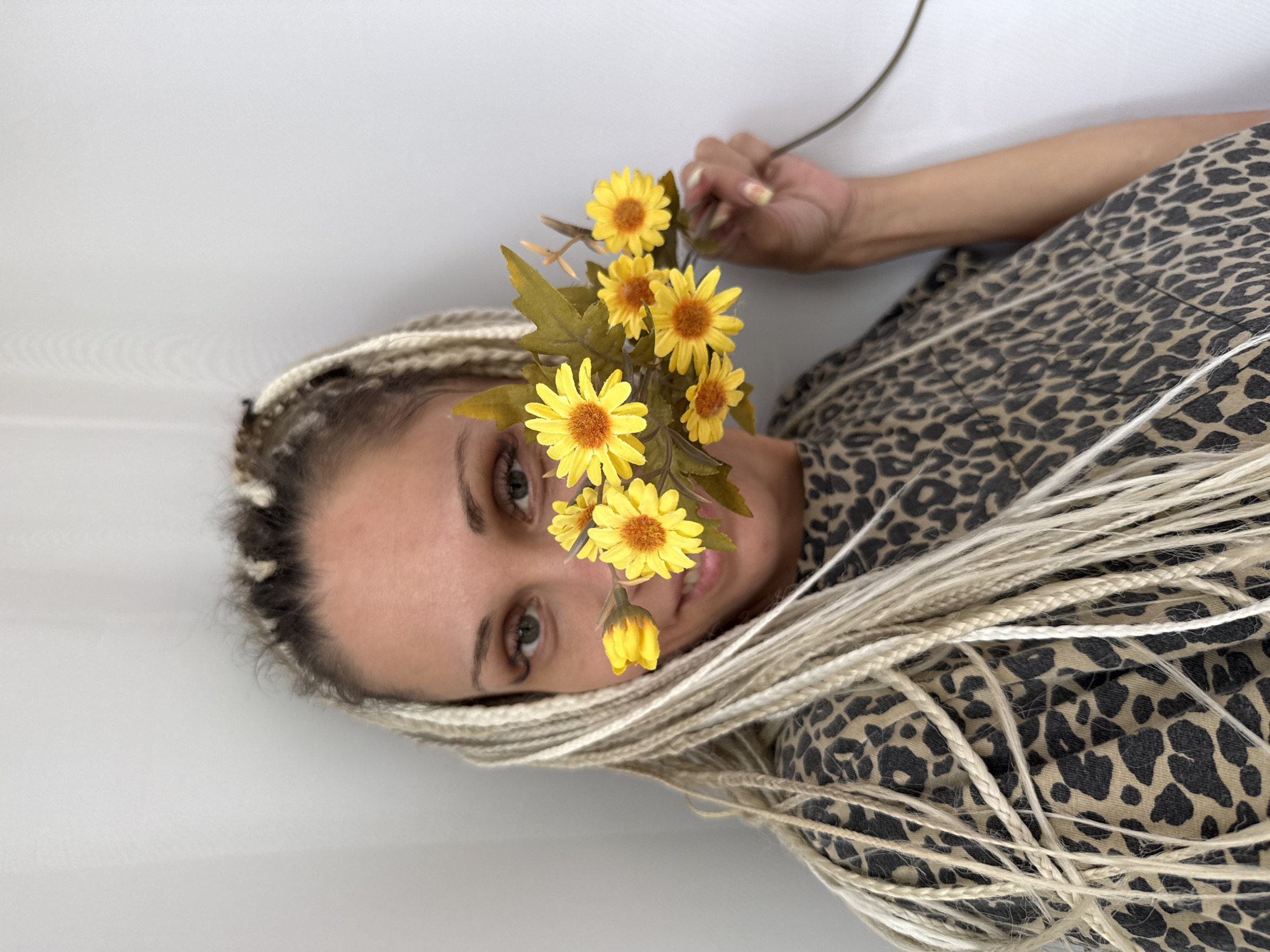 In Rusanovo, Russia, a woman with blonde dreadlocks and a leopard-print top is partially hidden behind a bouquet of yellow daisies. Her eyes, green, look directly at the camera, while her mouth is obscured by the flowers. The background is a plain white wall, creating a minimalist setting. The woman's right hand is visible, holding the stem of the daisy bouquet. Her nails are painted with a light yellow hue.