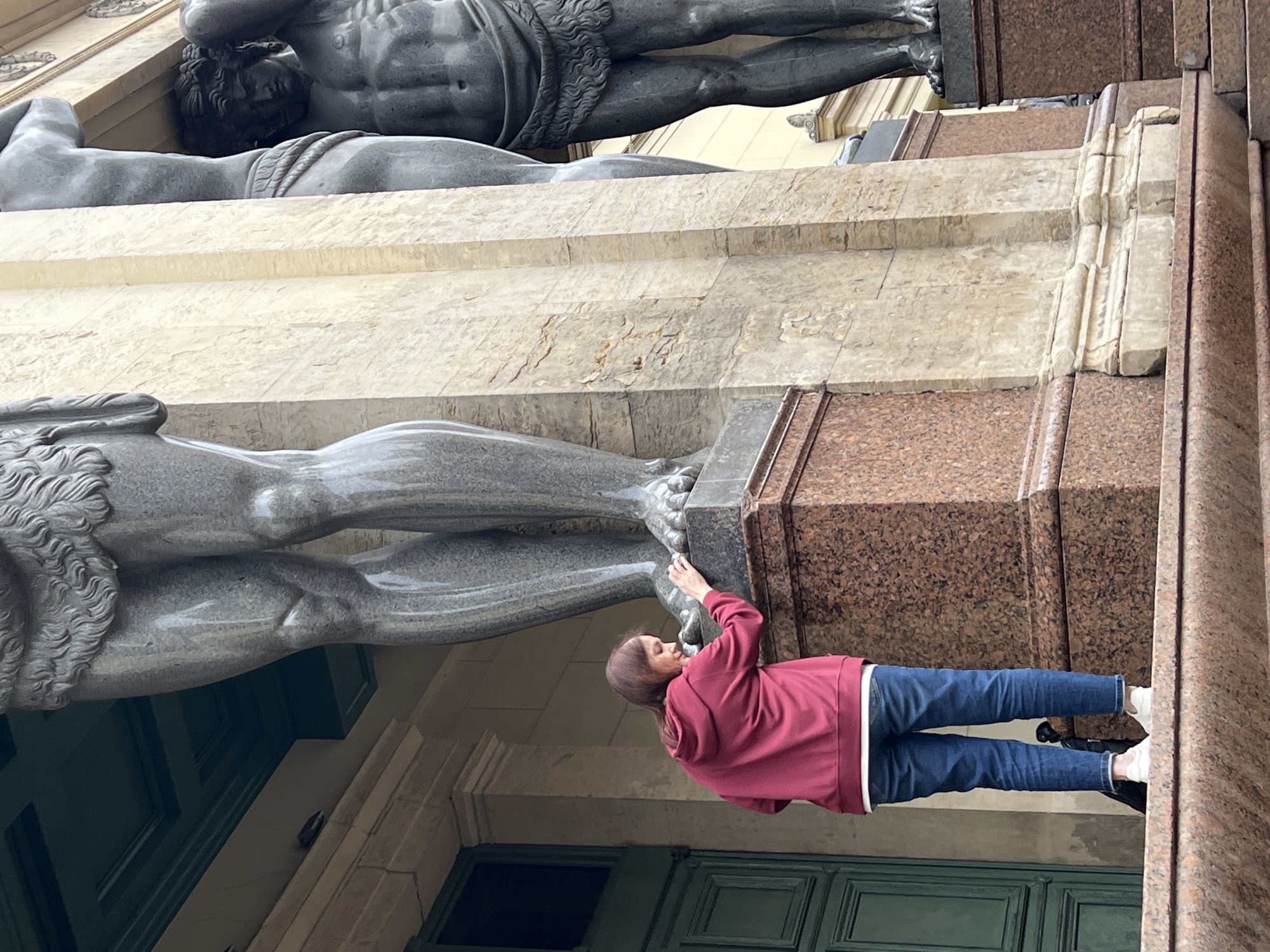 A woman in a maroon hoodie and blue jeans stands on a granite staircase, leaning towards a colossal stone statue of a man's legs. The statue is part of an architectural facade, likely a building in Saint Petersburg, Russia. The woman's right hand is touching the base of the statue. Behind her, a large green door is visible. To the right, another massive stone statue of a man is partially visible, supporting an unseen structure. The lighting suggests it is daytime, and the weather appears overcast.