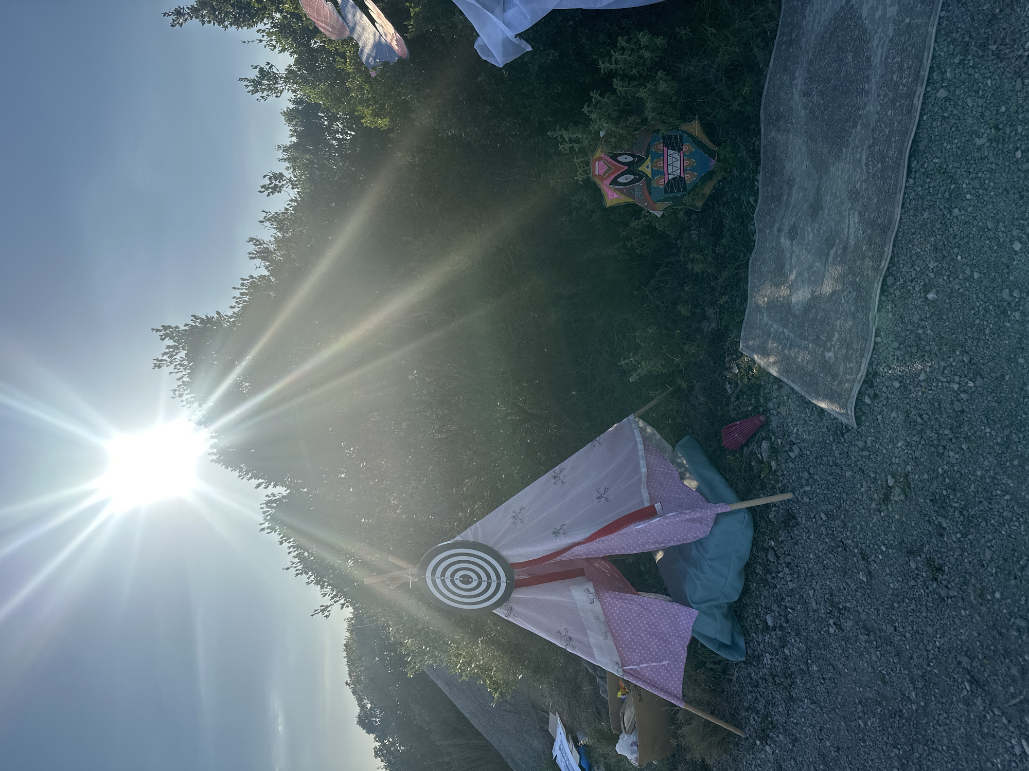 The image depicts an outdoor scene in Bogišići, Montenegro under bright sunlight. Various objects are laid on the ground next to vegetation and a gravel-covered area. Items include a decorated kite, a piece of fabric, and a target-like object attached to poles. The background includes trees and a clear sky obscured by the sun's glare. No people are visible.