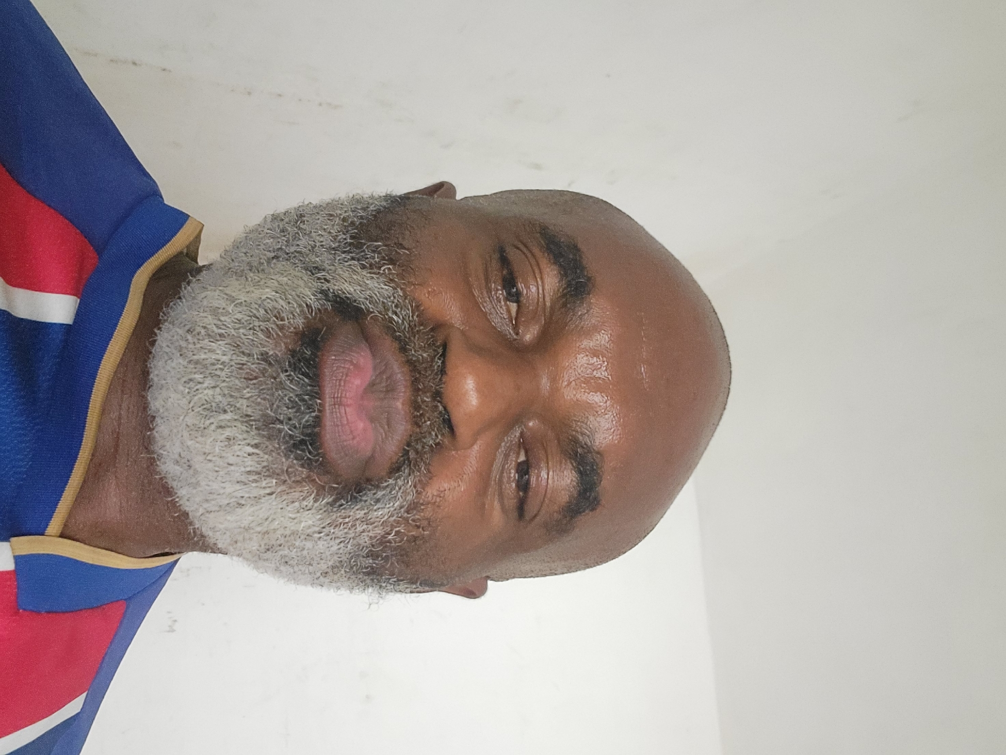 A close-up, indoor shot of a Black man with a bald head and a gray beard. He is wearing a shirt with blue, red, and white stripes. The background is a plain, white wall. The location is not discernible from the image.