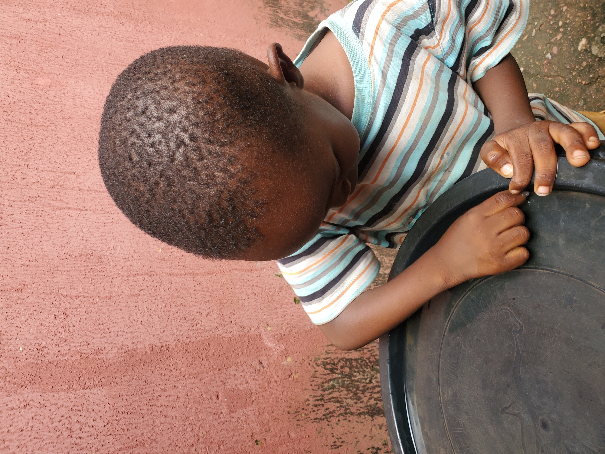 A close-up, high-angle shot shows the head and arm of a young Black child leaning over a black plastic container. The child is wearing a horizontally striped t-shirt with colors of teal, white, orange, and black. The child's short, dark hair is visible, as is their ear. Their right arm rests on the rim of the container, with their fingers spread slightly. Their left hand is also visible, resting on the container. The ground beneath the child appears to be made of reddish-pink concrete or stucco, with some patches of darker, possibly wet, material. The overall lighting suggests daytime, though no specific time can be determined. The scene appears to be outdoors in an urban or residential setting. Visible text on the container includes "PLASTICS". The context of Jos, Nigeria, implies this scene is set within that geographical location.