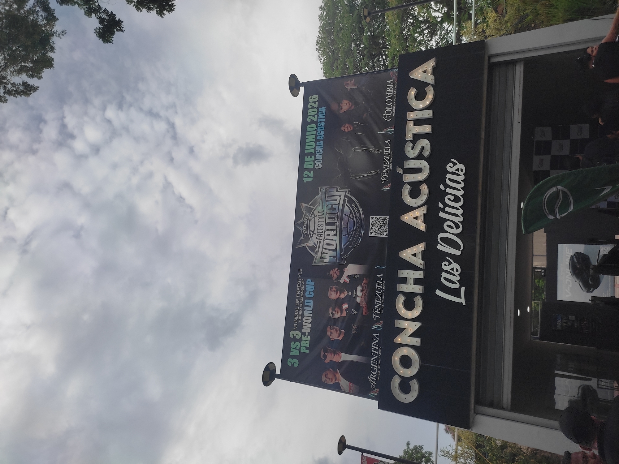 A wide shot captures the exterior of a venue in Maracay, Venezuela, under a cloudy sky. Dominating the scene is a large black banner advertising a "3 VS 3 MUNDIAL DE FREESTYLE PRE-WORLD CUP" event. The banner features images of several young men, likely participants, and displays "ARGENTINA," "VENEZUELA," and "COLOMBIA" with team names listed below. The date "12 DE JUNIO 2026" is prominently displayed, alongside the venue name "CONCHA ACÚSTICA." Below the banner, the venue's entrance is visible, with large, illuminated letters spelling out "CONCHA ACÚSTICA" above "Las Delicias." Several individuals, some wearing dark clothing and caps, are partially visible near the entrance, with some appearing to be patrons or staff. A green banner or flag is positioned to the left of the entrance. The overall atmosphere appears to be that of an outdoor event venue in a tropical or subtropical environment, given the lush green foliage visible in the upper right corner.