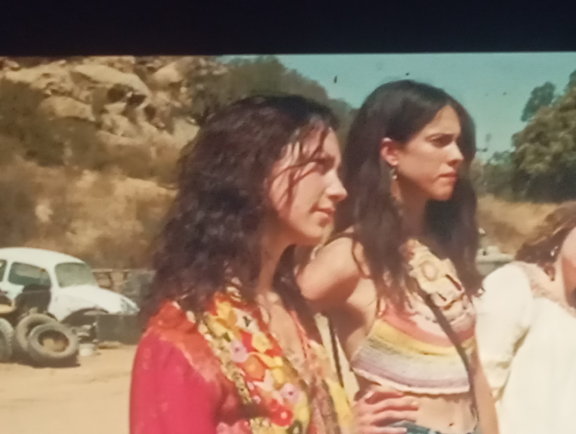 The image depicts two young women standing outdoors. The woman on the left has dark, curly hair and is wearing a vibrant, floral-patterned shirt or scarf. The woman on the right has long, dark hair and is wearing a crocheted top with rainbow stripes. In the background, there is a derelict white car and several tires. The environment appears to be arid or semi-arid, with dry vegetation and rocky terrain. The sky is a pale blue. The lighting suggests it is daytime.